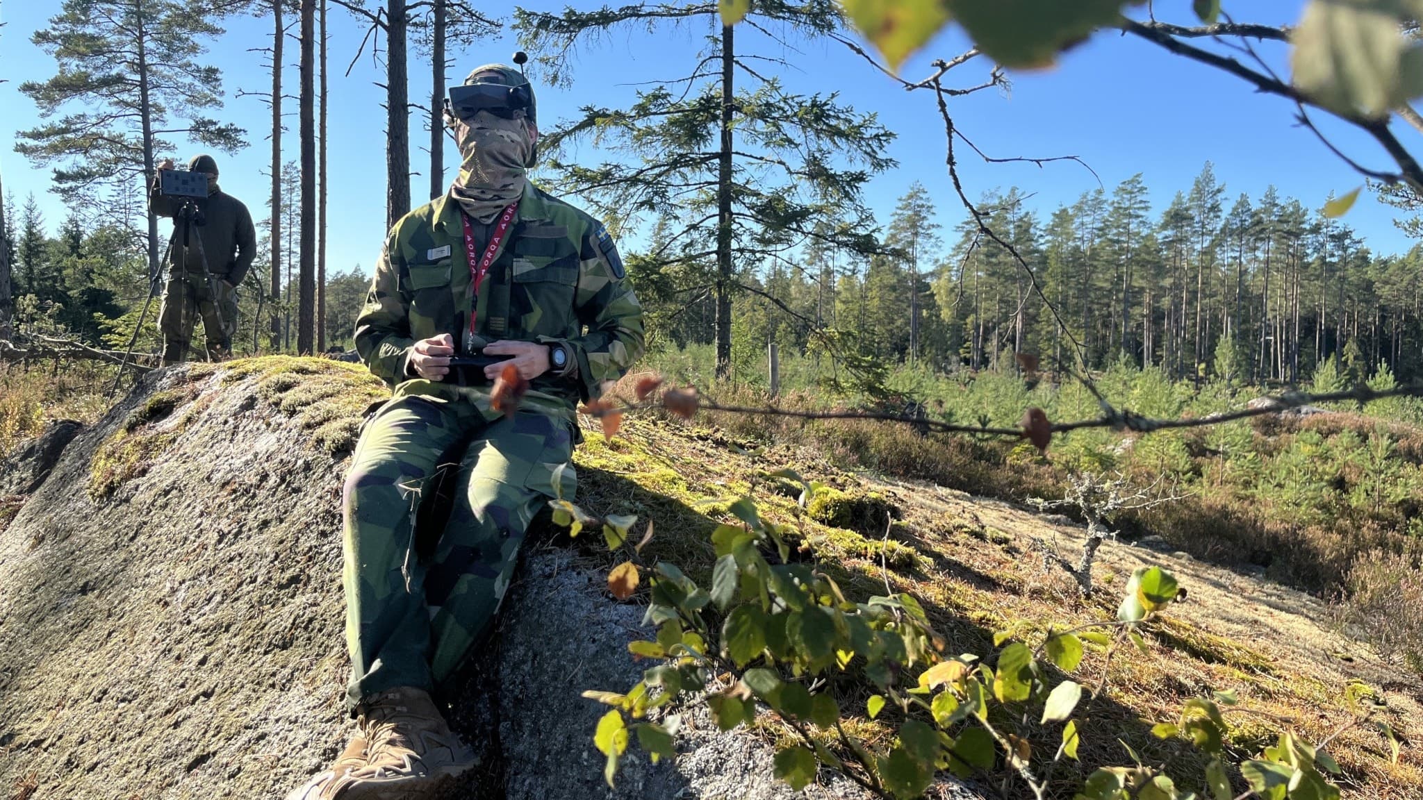 En jägarsoldat i militäruniform sitter på en klippa i skogen med en drönarkontroll i handen. I bakgrunden syns instruktören Johan stående bland träd.