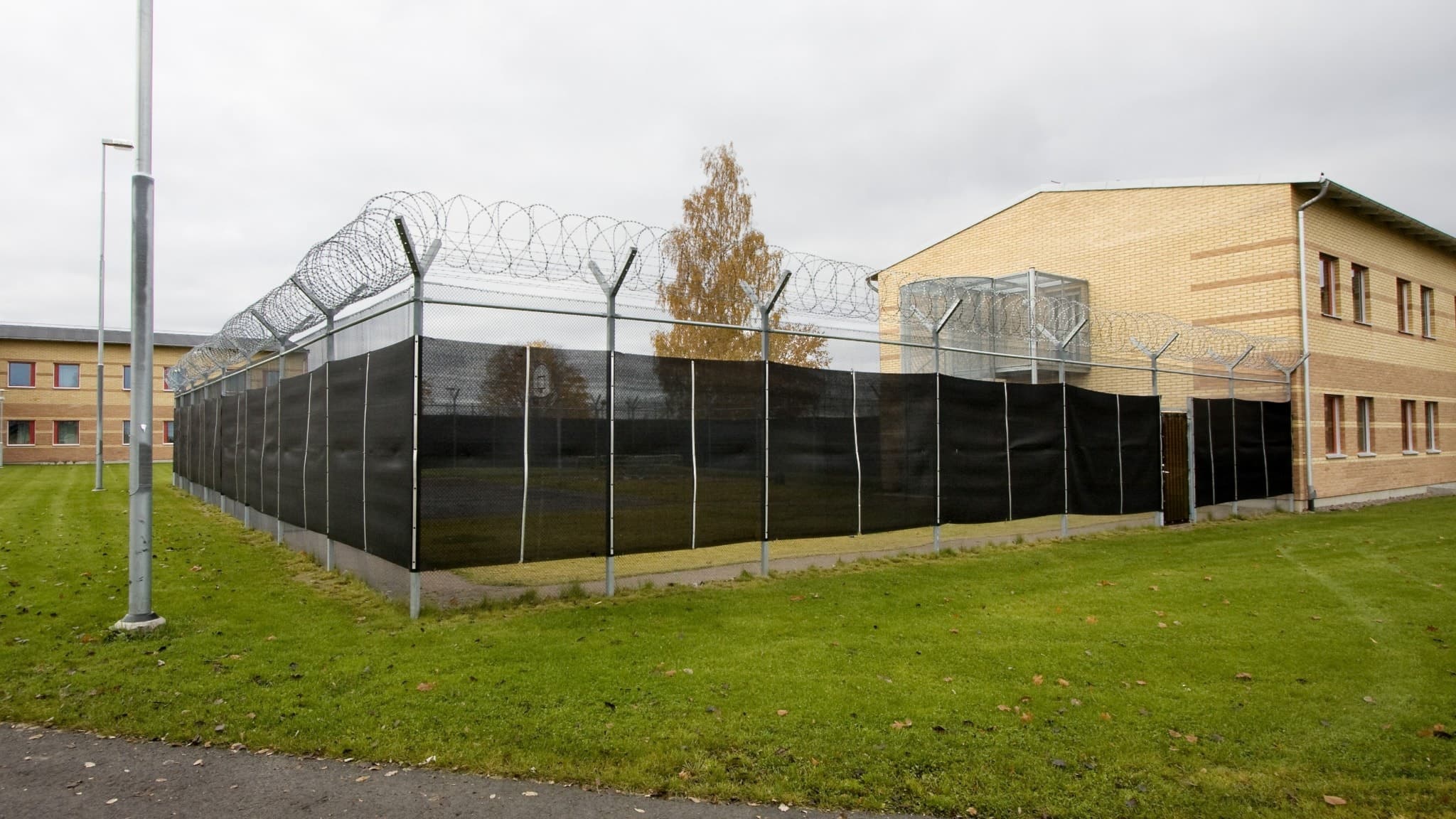 Bild på anstalten Skänninge med gula fasader, höga staket och taggtråd som omgärdar byggnaden. Grön gräsmatta syns i förgrunden under en gråmulen himmel.