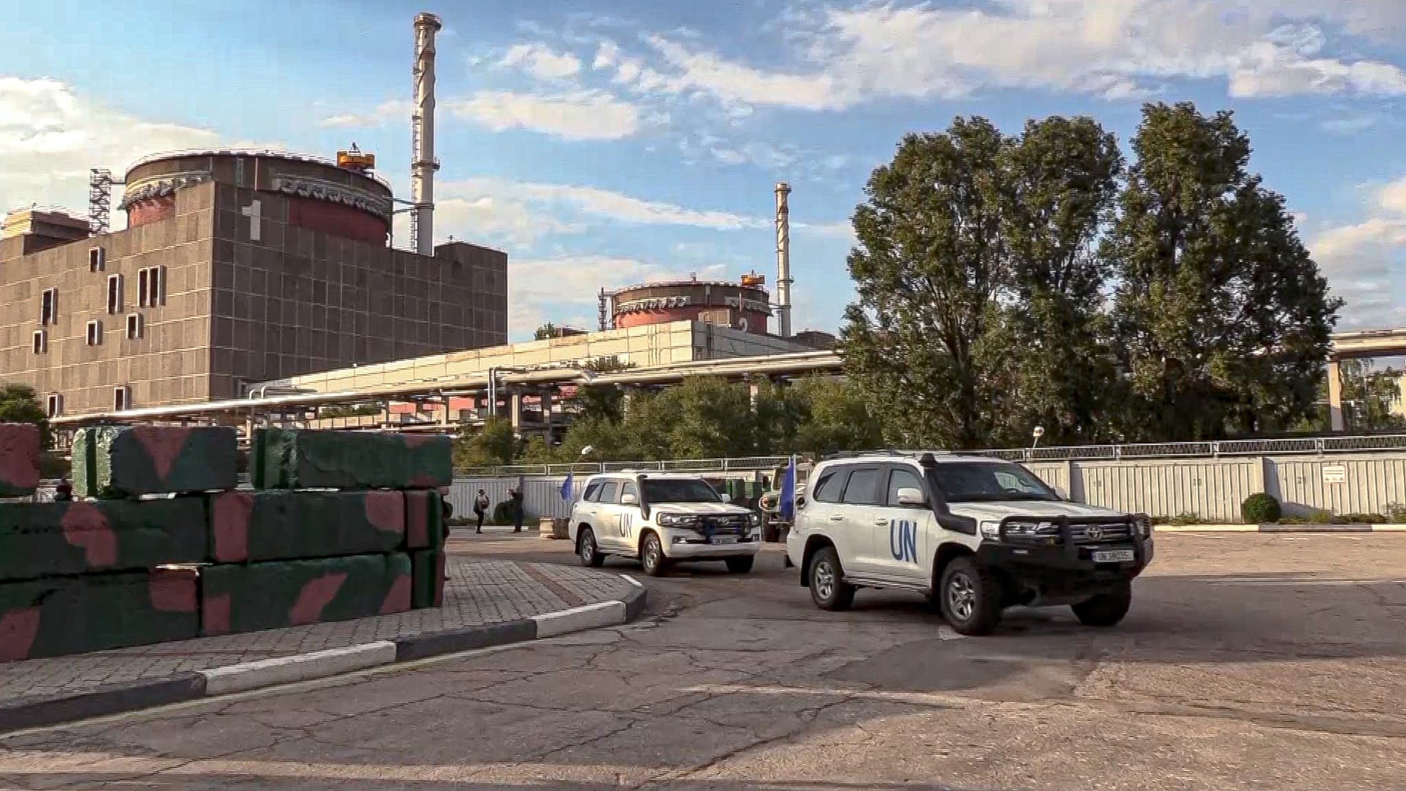 Bilar från FN:s atomenergiorgan anländer till kärnkraftverket Zaporizjzja i Ukraina. Byggnader och säkerhetsbarriärer syns i bakgrunden under en solig dag.