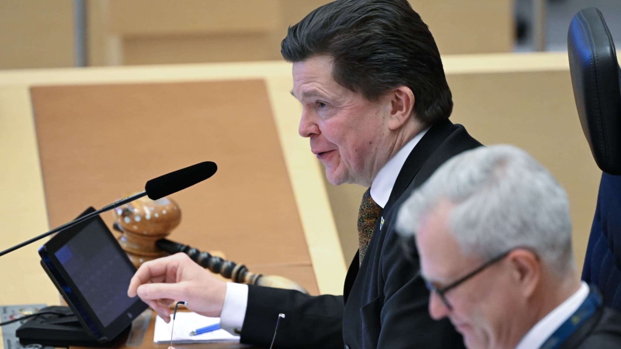 Riksdagens talman Andreas Norlén sitter vid ett podium under en partiledardebatt i riksdagen. Han är klädd i kostym och har dokument framför sig, med mikrofoner och en skärm på bordet.