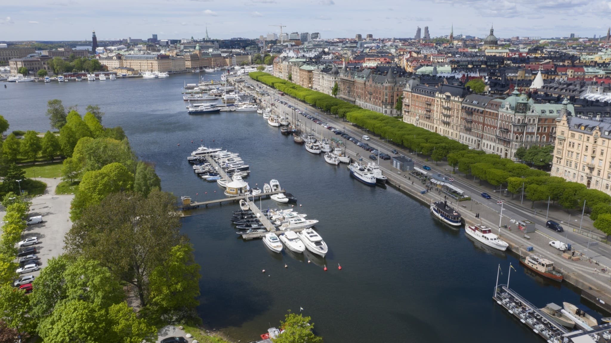 Sommarvy över Strandvägen i Stockholm med båtar längs kajen, grönskande träd och Östermalms karakteristiska byggnader i bakgrunden. En populär plats för promenader och båtliv.