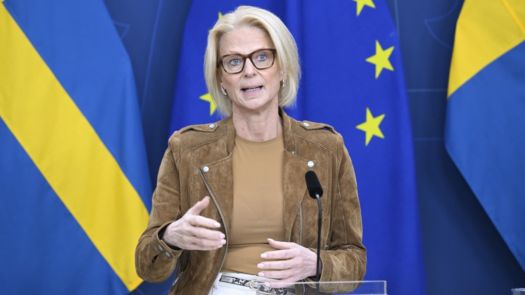 Finansminister Elisabeth Svantesson (M) talar vid en pressträff om kärnkraft, med svenska och EU-flaggor i bakgrunden. Hon står vid ett podium och gestikulerar under presentationen.