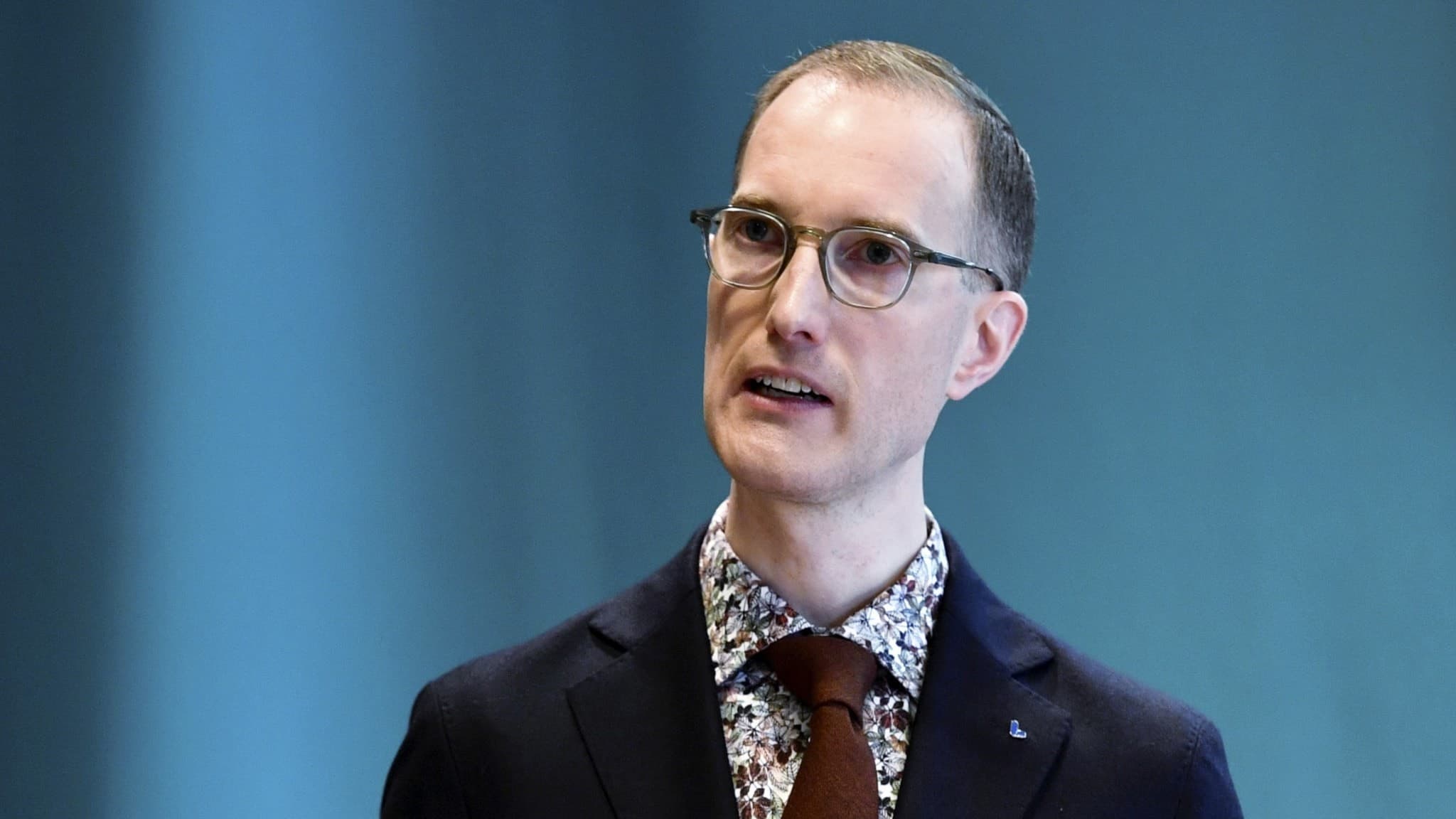 En man i kostym talar vid ett podium med en mikrofon framför sig. Bakgrunden är blå och ett glas vatten står på podiet. Fokus ligger på formell presentation i en professionell miljö.