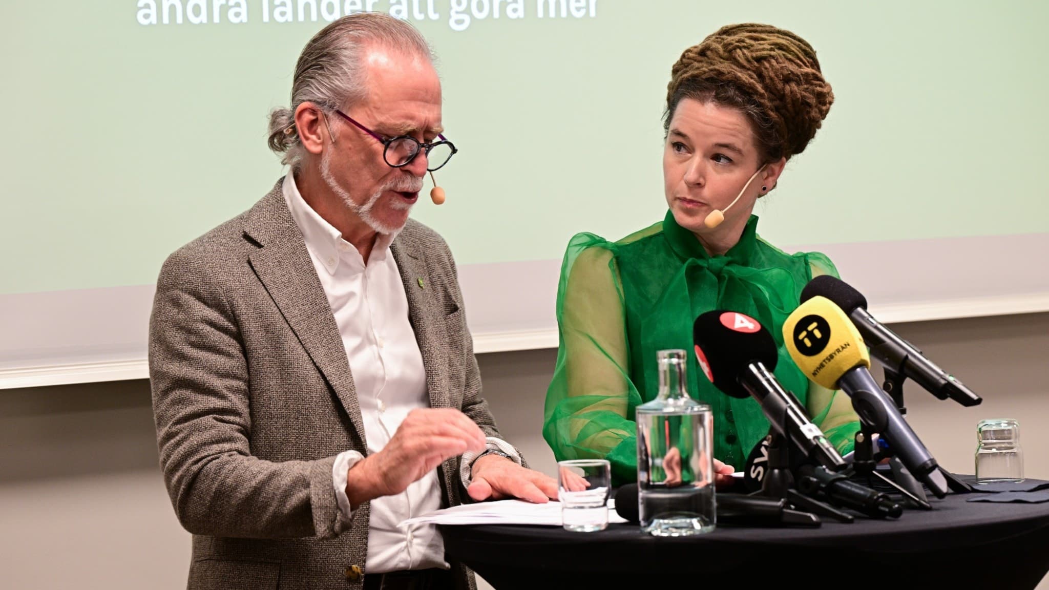 Miljöpartiets språkrör Daniel Helldén och Amanda Lind står vid ett podium med mikrofoner under en pressträff på Miljöpartiets kongress i Västerås. En vattenkaraff och glas syns på bordet.