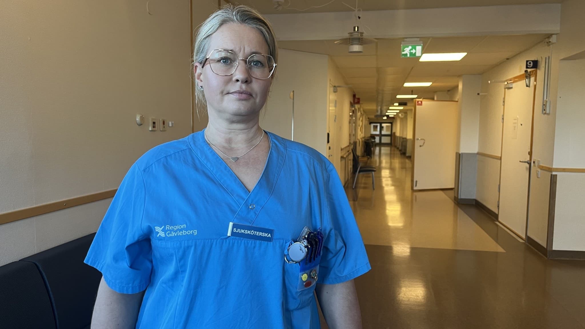 Sjuksköterskan Linda Gillberg på Familjeläkarjouren i Gävle står i en tom sjukhuskorridor. Hon bär blå arbetskläder och glasögon.