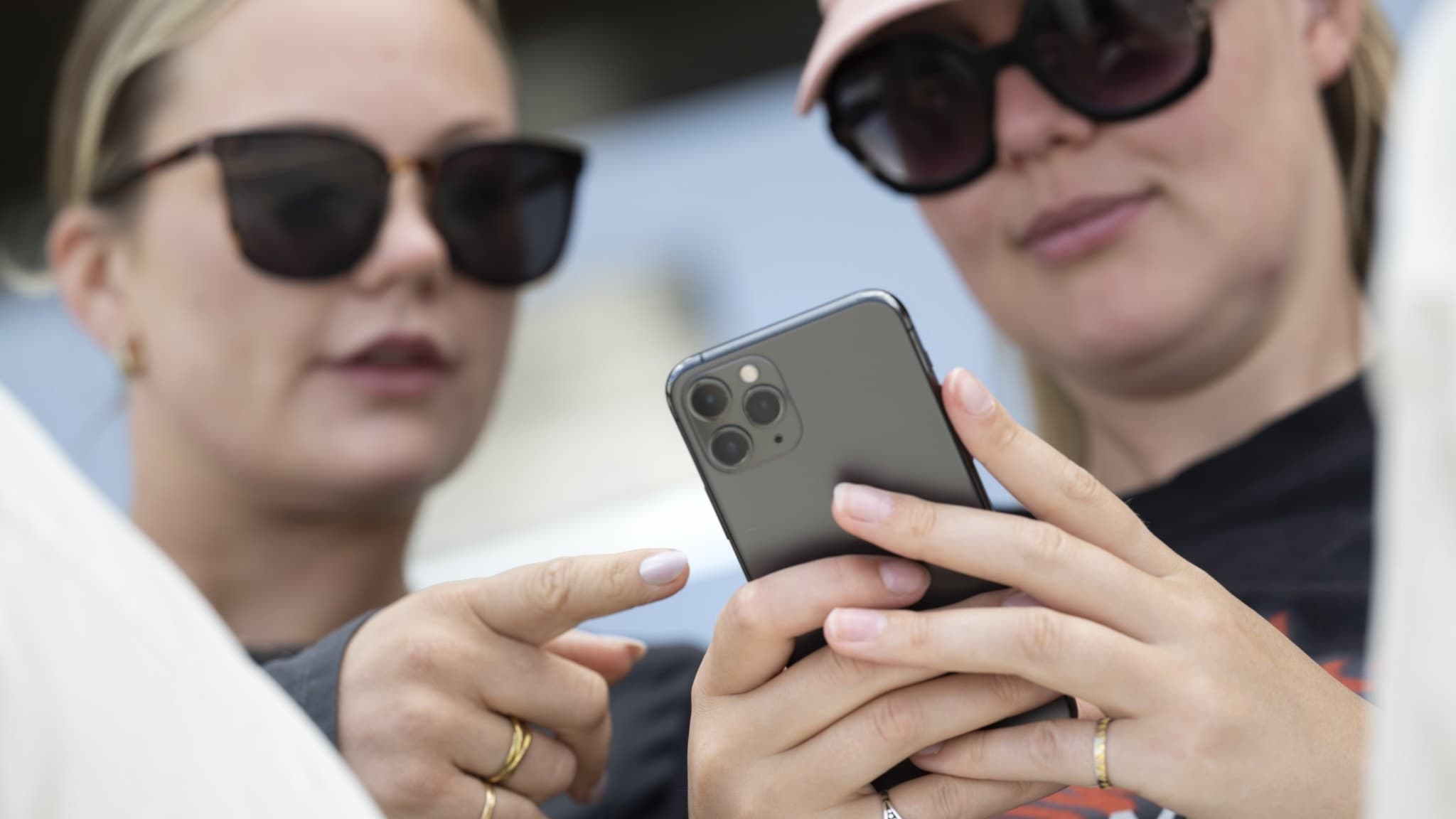 Två kvinnor i Stockholm håller en modern smartphone och använder sociala medier, med fokus på skärmen. De har solglasögon och ser ut att vara engagerade i vad som visas.