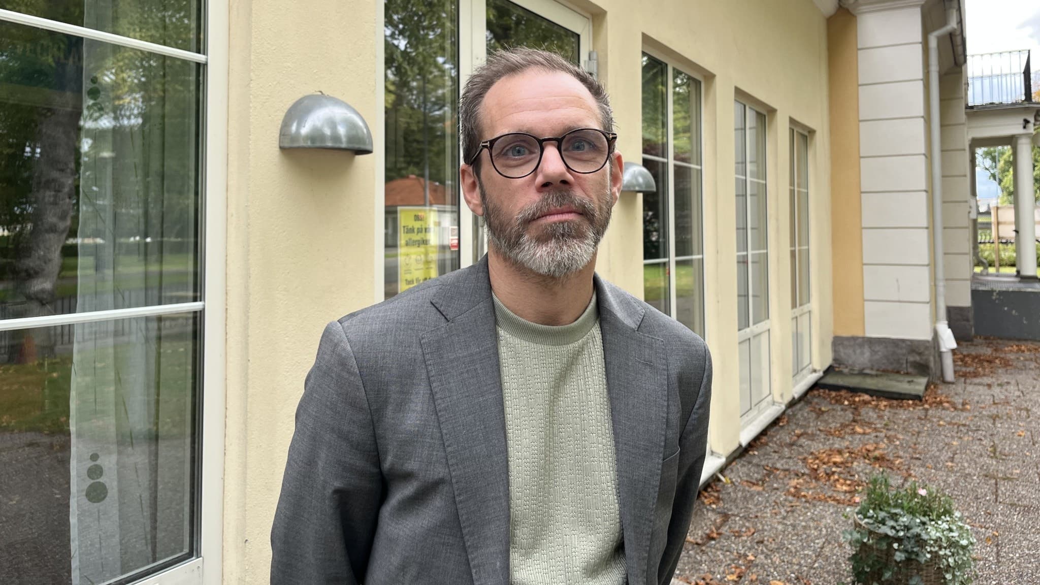 En man i kavaj står framför en gul byggnad med stora glasfönster.