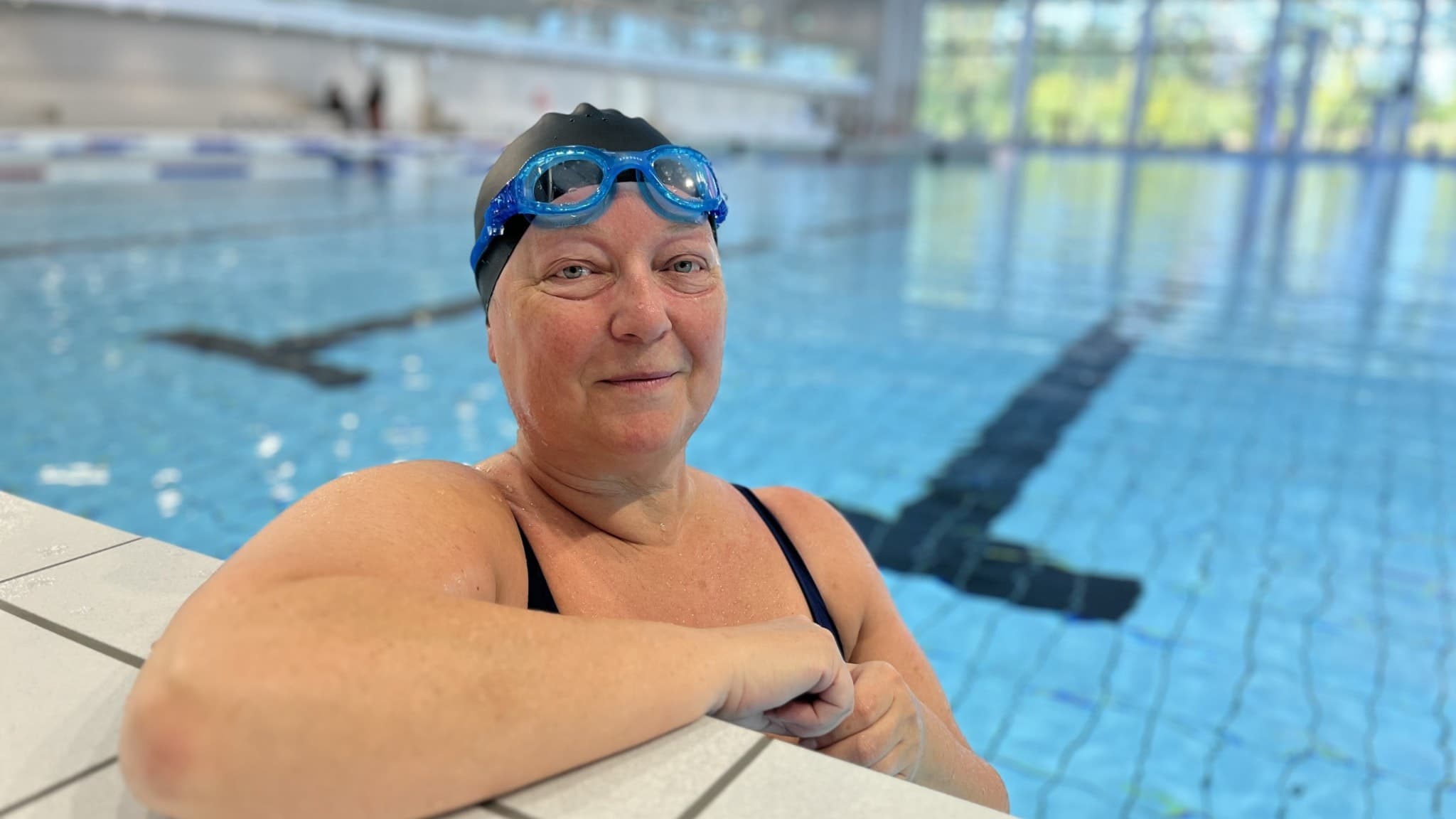 Maria Nilsson, 17, står vid kanten av en stor inomhuspool med klart blått vatten. Hon bär en svart baddräkt och simglasögon. Bakgrunden visar stora fönster och simbanor.