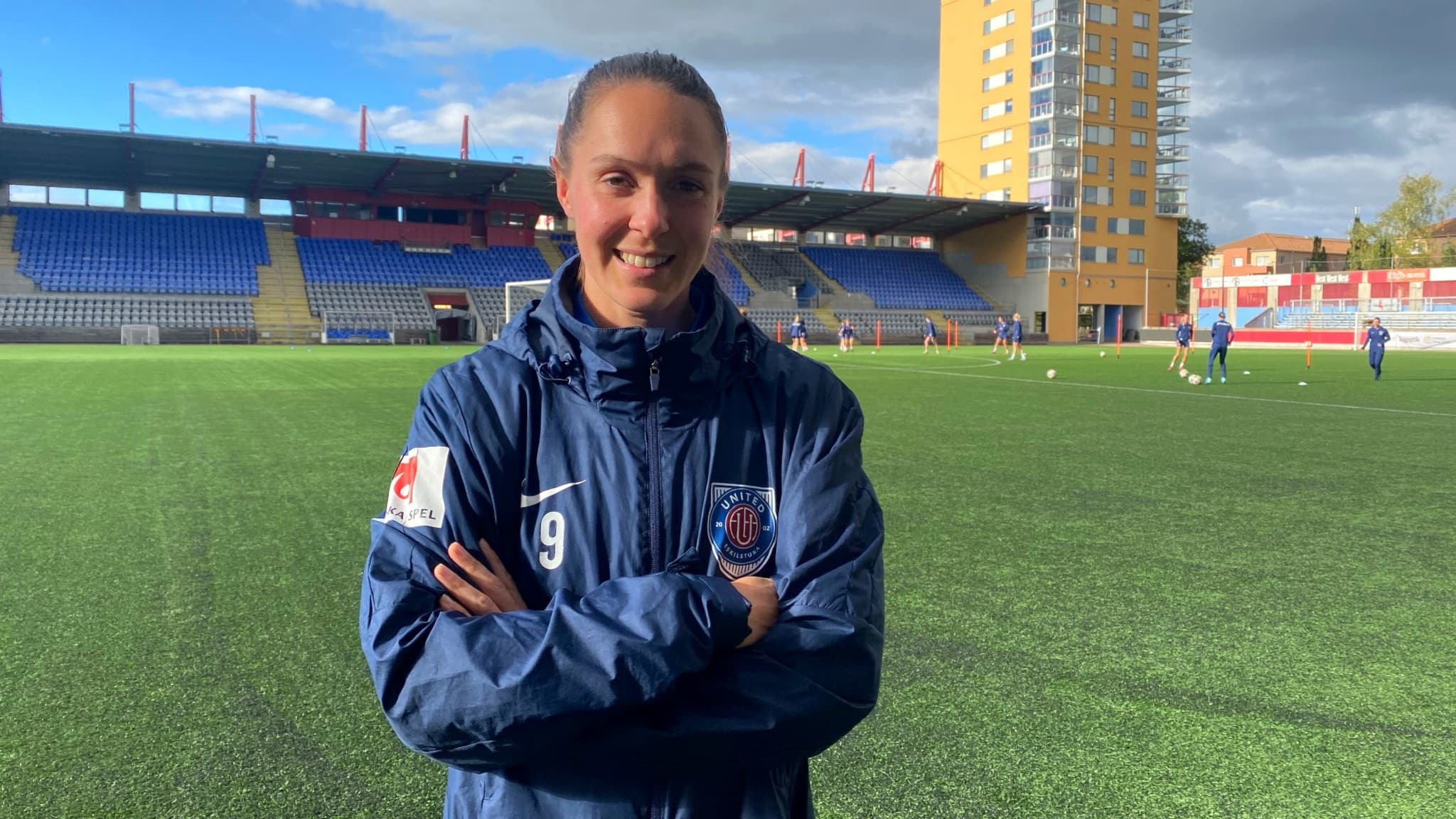 Felicia Rogic står på en fotbollsplan iklädd en blå träningsjacka med korsade armar. I bakgrunden syns en läktare, en grön konstgräsplan och en gul byggnad.