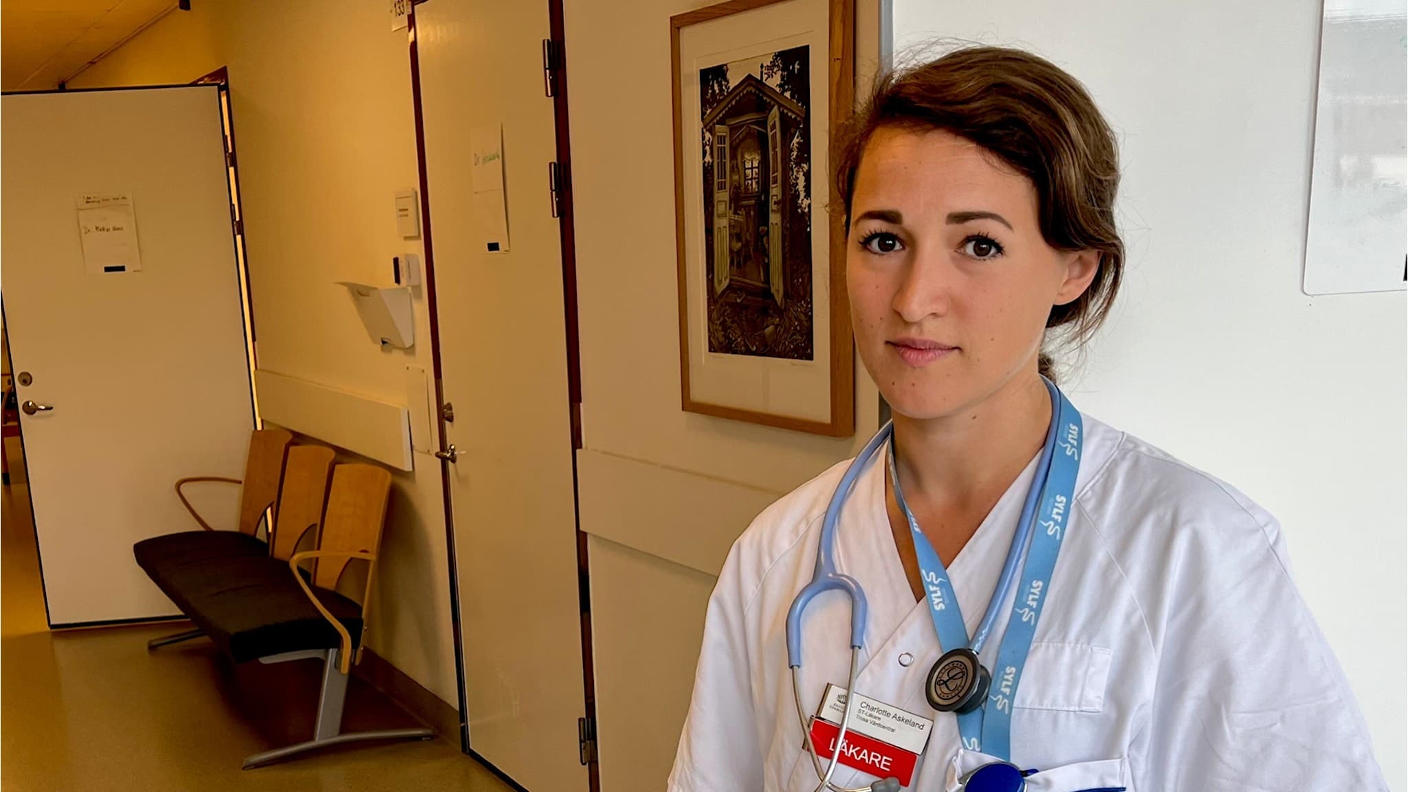 Charlotte Askeland, ST-läkare, står i en sjukhuskorridor iklädd vit läkarrock med ett stetoskop runt halsen. Korridoren är ljus med öppna dörrar och en bänk längs väggen.