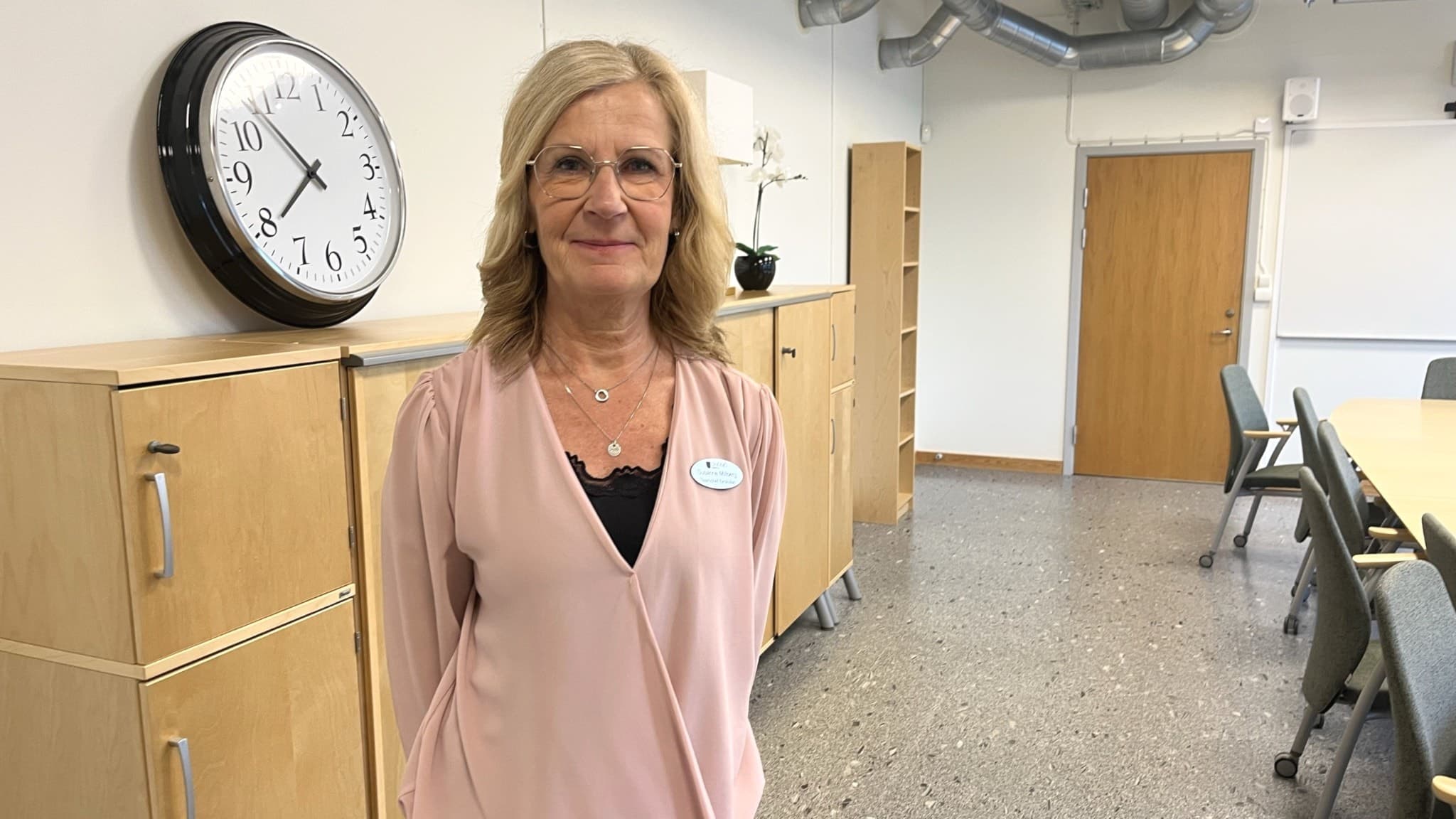 Susanne Millberg står i ett kontorsrum/klassrum med ljusa väggar, en klocka på väggen och hyllor i trä. 