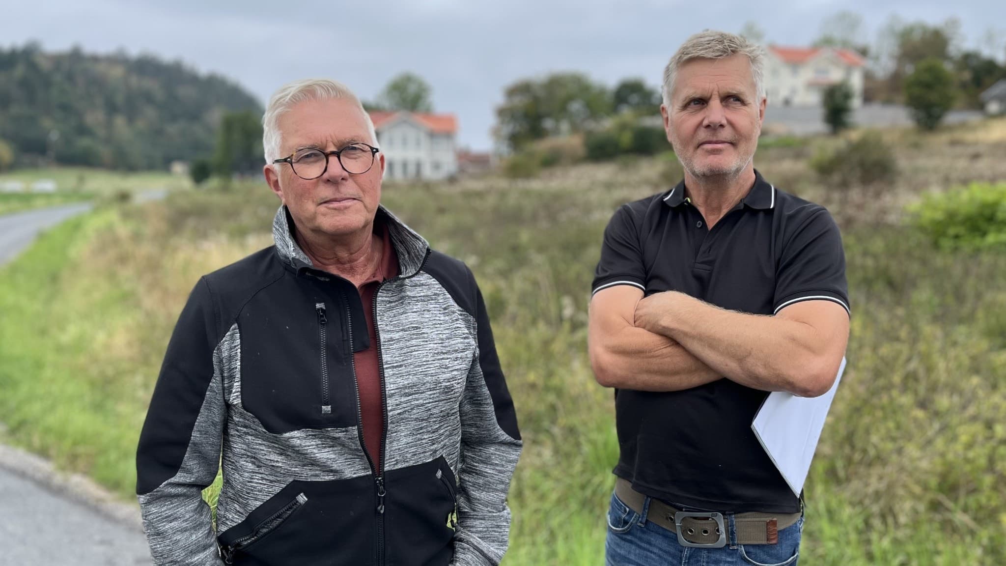 Peter Karlsson och Jonas Brusbäck står vid en landsväg omgivna av grönska och hus i bakgrunden. Karlsson bär en mönstrad jacka medan Brusbäck är klädd i en svart pikétröja.