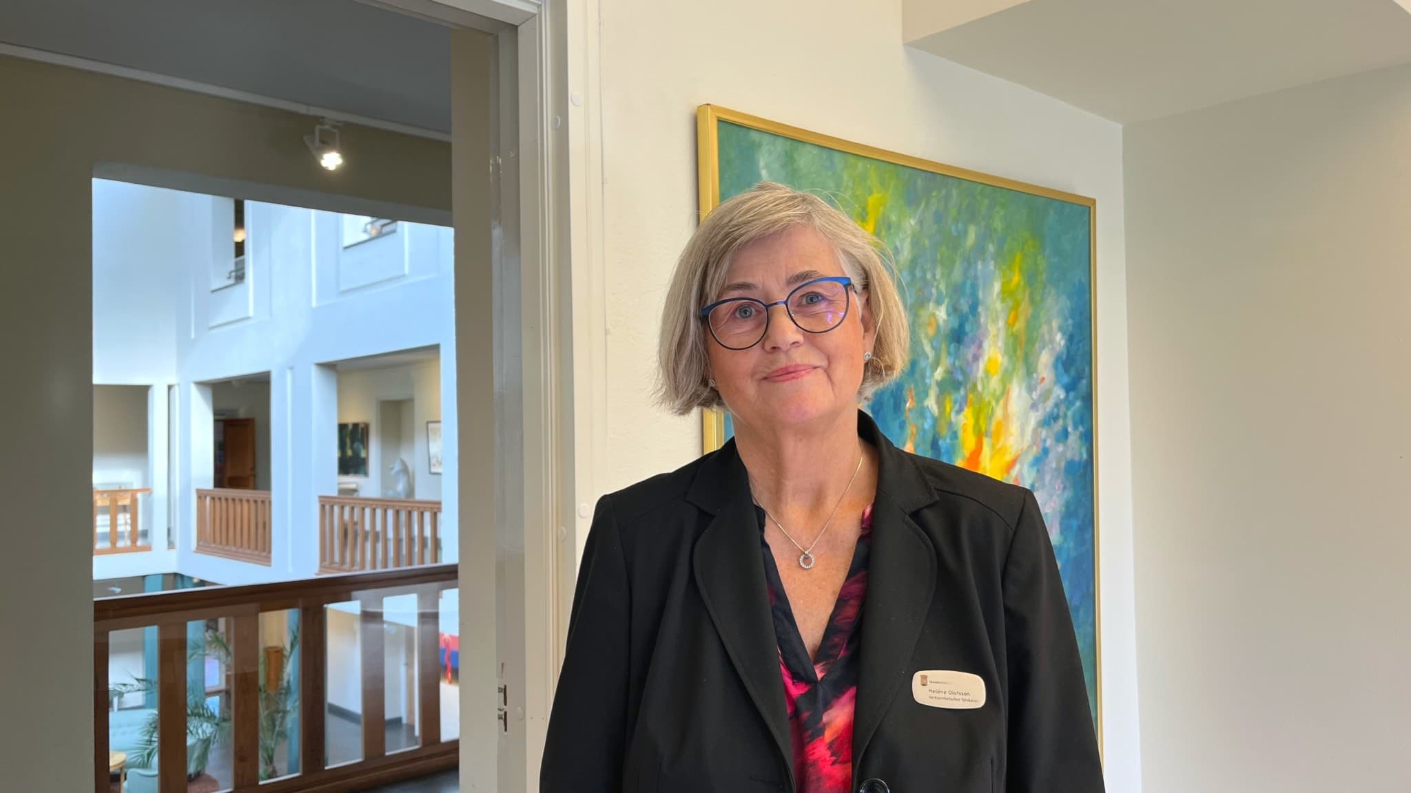 Förskolechef Helene Olofsson står inomhus vid en ljus vägg med en färgglad tavla i bakgrunden. Hon har en mörk kavaj och ett namnkort på bröstet. Miljön visar en modern byggnad.
