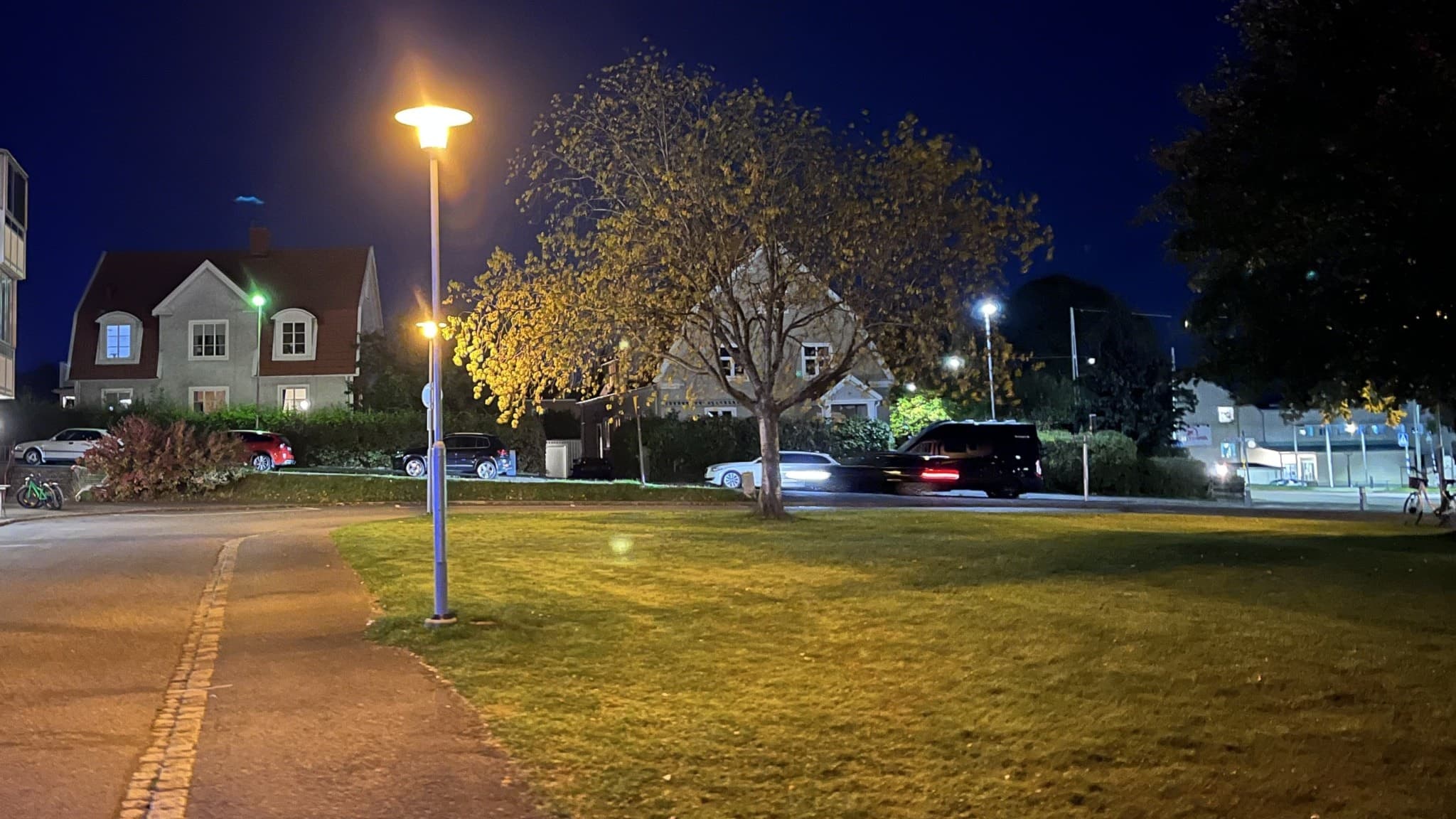 Rosenlundstorg på kvällen med en upplyst gatlykta, parkerade bilar och ett avspärrningsband på gräsytan. En person har förts till sjukhus efter att ha skadats med ett vasst tillhygge.