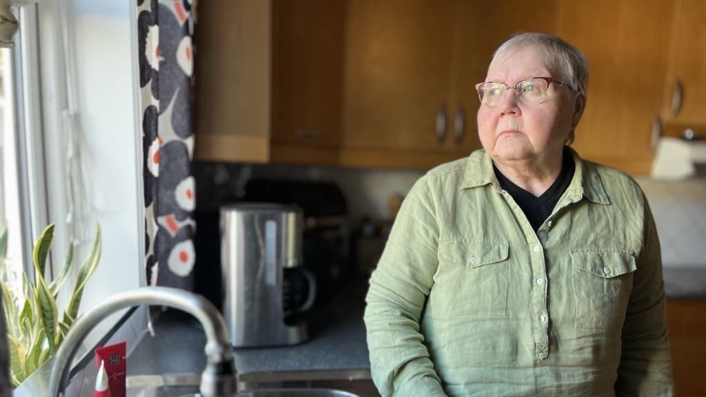 Ulla Svensson står i ett kök med träskåp och en diskho i förgrunden. En vattenkokare syns på bänken bredvid henne, och köksfönstret släpper in dagsljus.