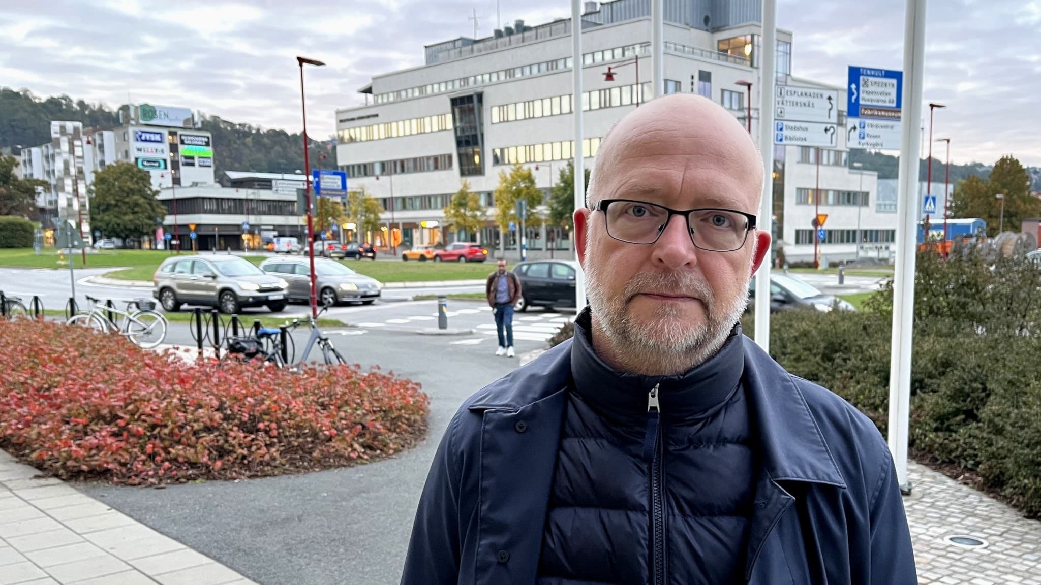 Carsten Wulf står utomhus framför en modern stadsmiljö med byggnader, flaggstänger och buskar i bakgrunden. Himlen är molnig och flera personer rör sig i området.