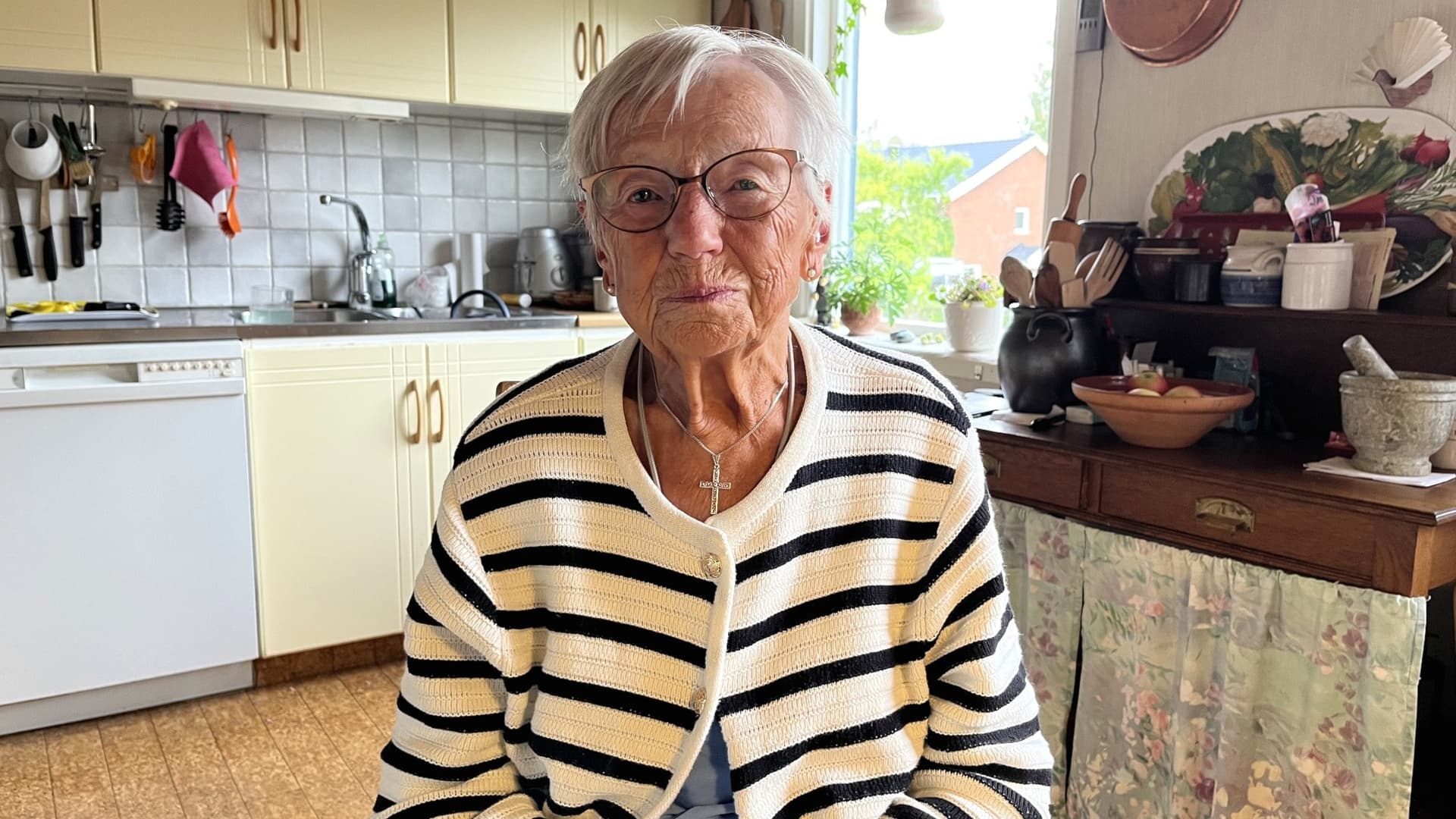 Mirjam Ring sitter i ett lantligt kök med ljusa skåp och en rutigt klädd köksbänk i bakgrunden. Hon bär en randig tröja och ett halsband.