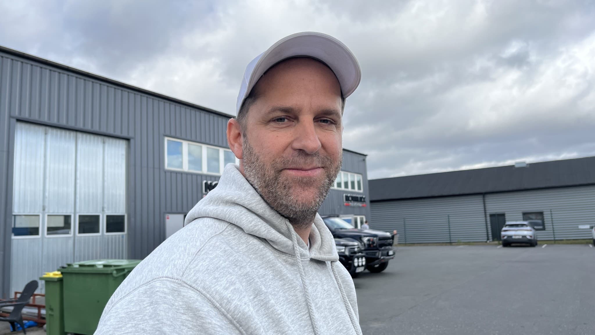 En person i grå hoodie och vit keps står utomhus framför en industribyggnad med grå fasad. 