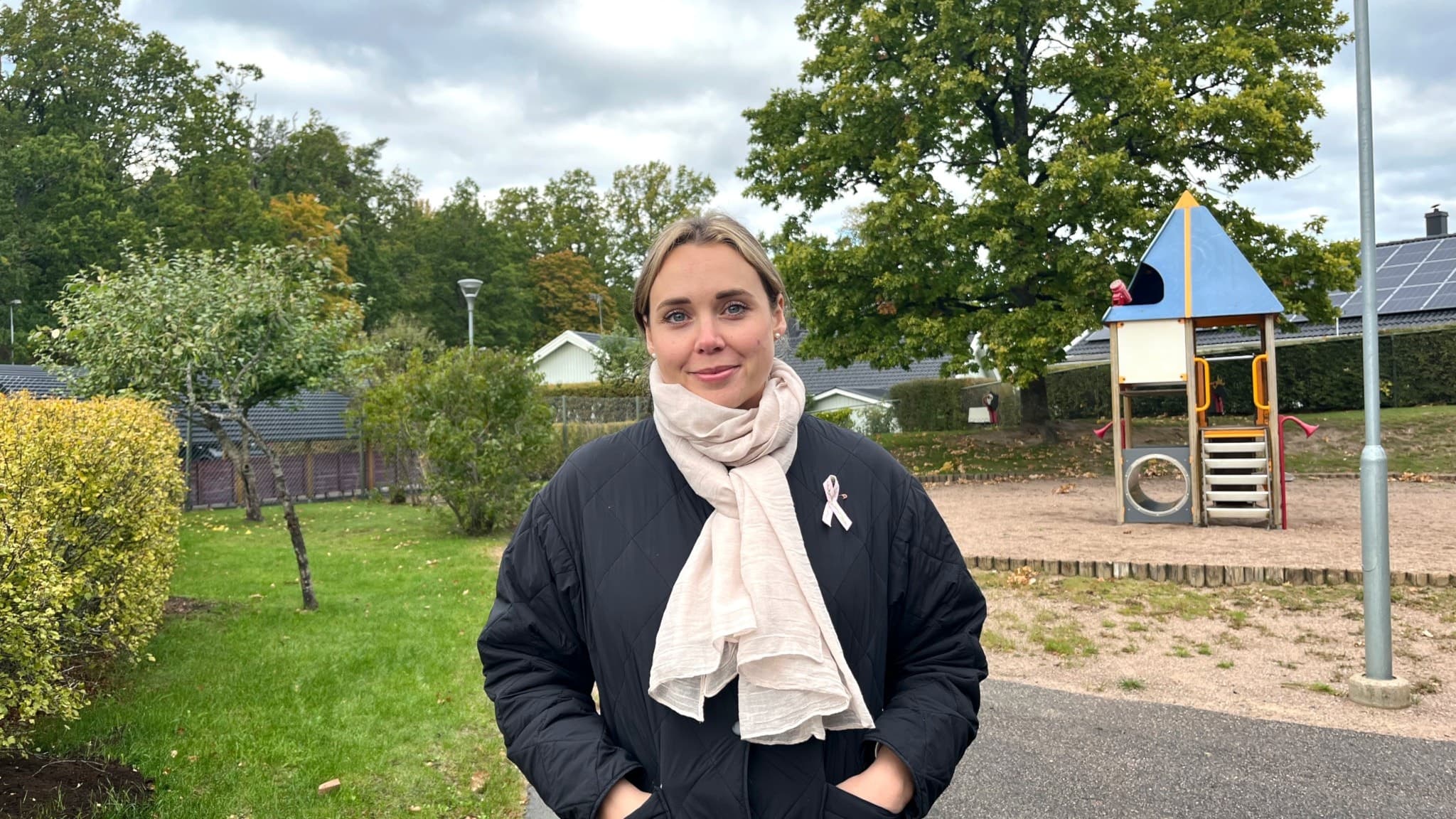 Julia Tjäder Millestad står utomhus i en park med en lekplats i bakgrunden. Hon bär en mörk jacka och en ljus halsduk. Grönska och molnig himmel syns i bakgrunden.