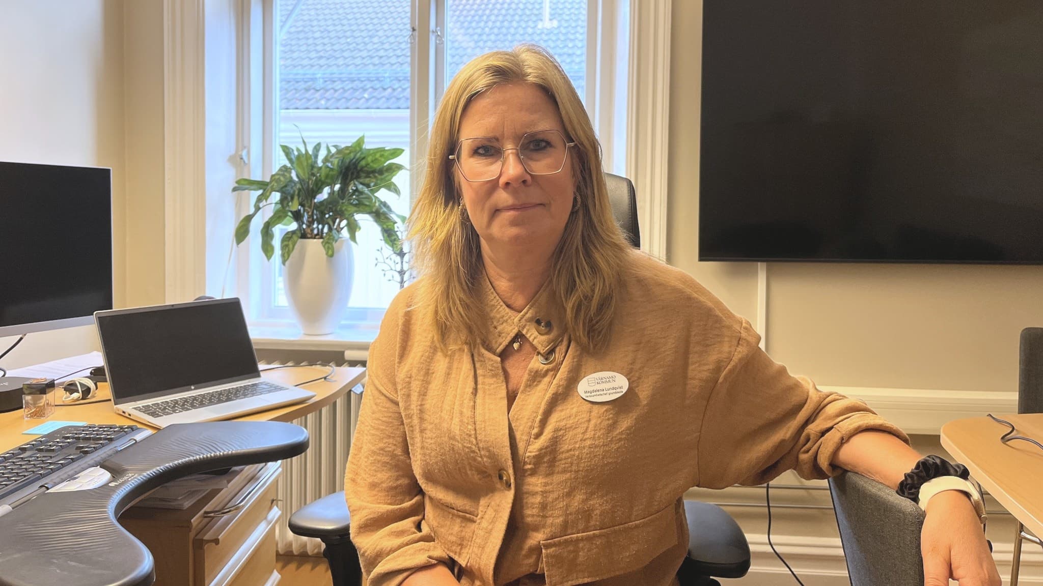 En kvinna som tittar in i kameran när hon sitter på sitt kontor. Bakom henne är det en kontorsmiljö av datorer, en tv samt en växt i fönstret.