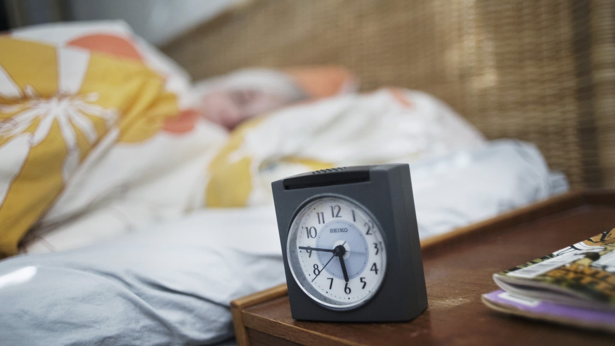 En kvinna sover i en säng med ett färgglatt täcke. I förgrunden syns en klassisk väckarklocka på ett nattduksbord, bredvid ligger böcker. Fokus ligger på klockan som visar morgontid.