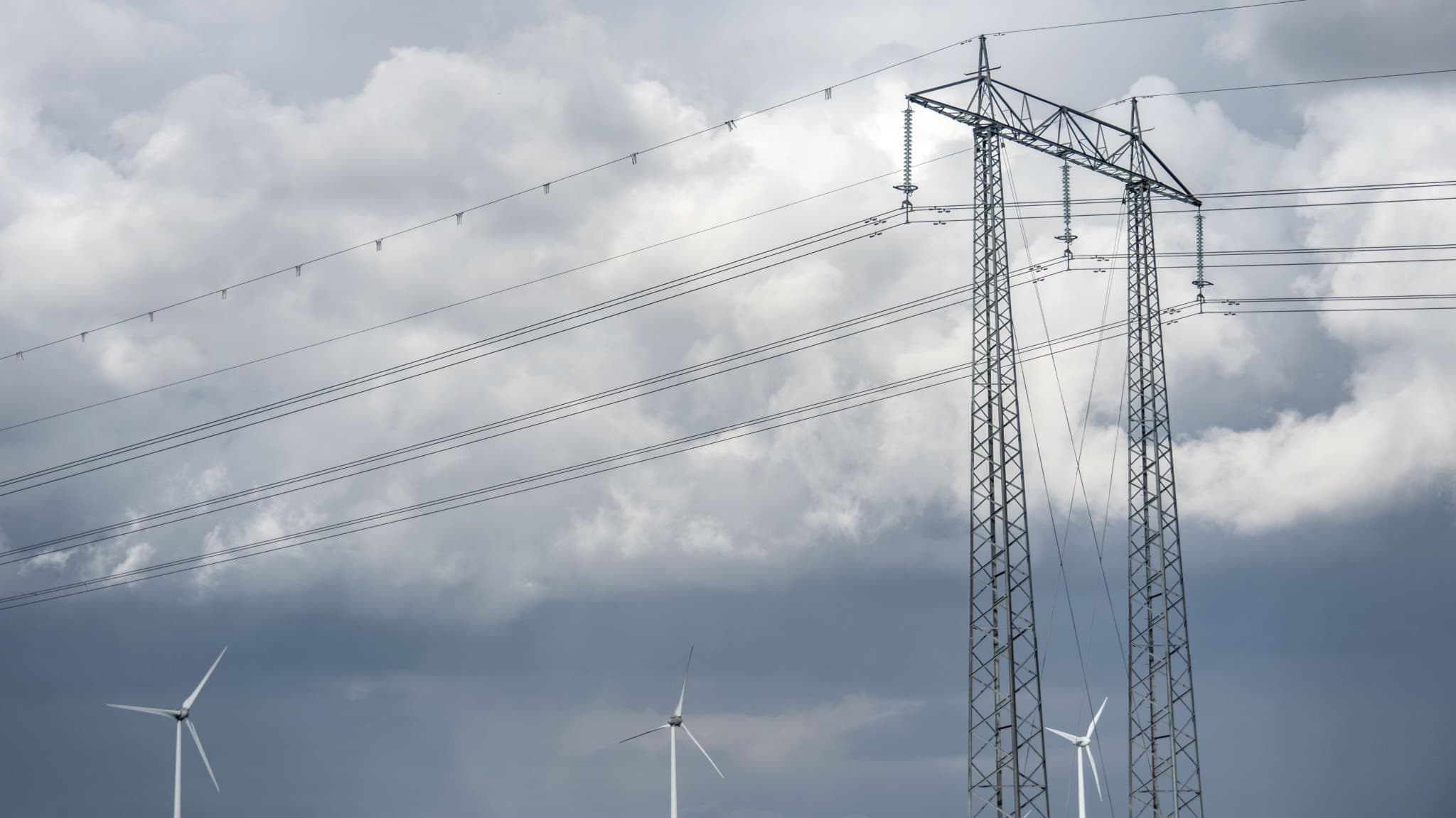 Mörka moln tornar upp sig bakom högspänningsledningar utanför Lund, med vindkraftverk som snurrar långsamt i svag vind på horisonten. En dramatisk himmel skapar en dyster atmosfär.