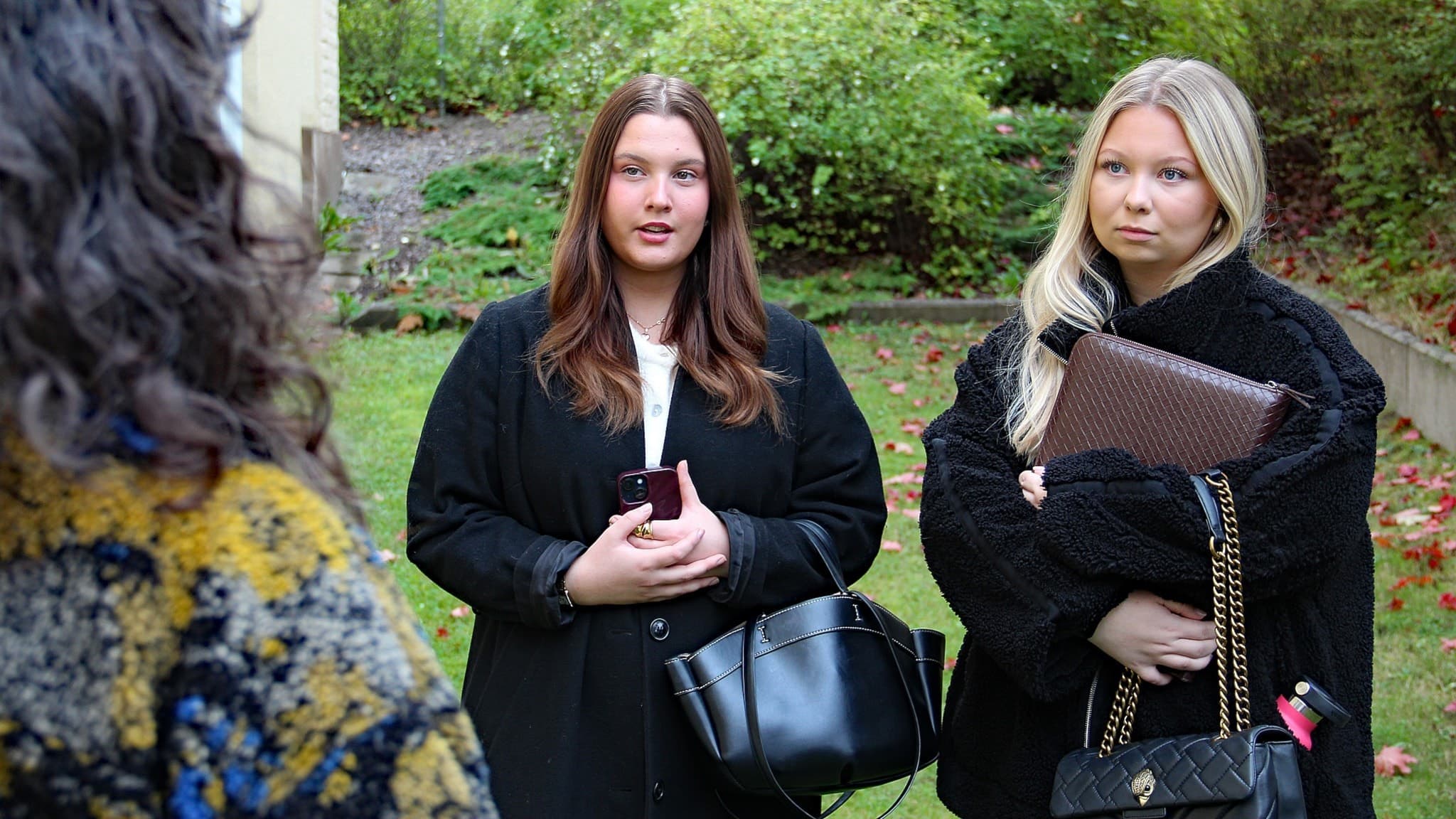 Emma Strömberg och Hella Jansson står utomhus i en trädgård, båda klädda i mörka kläder och håller handväskor. En tredje person syns bakifrån i förgrunden.