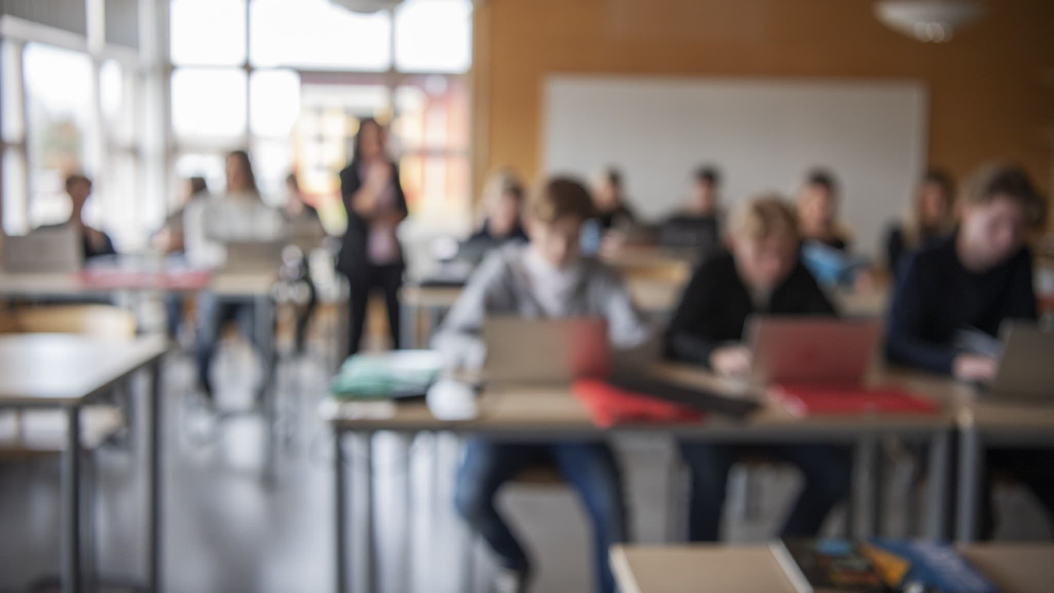 Elever sitter vid skolbänkar med bärbara datorer under en lektion i ett klassrum i Västra Götaland, Sverige. En lärare står längst fram i rummet nära en vit skrivtavla.