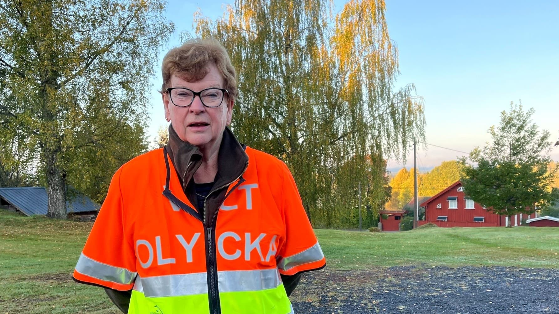 Lena Jansson eftersöksjägare, står utomhus i en reflexväst med texten 'Olycka' på. I bakgrunden syns en gårdsmiljö med träd och röda byggnader i morgonljus.