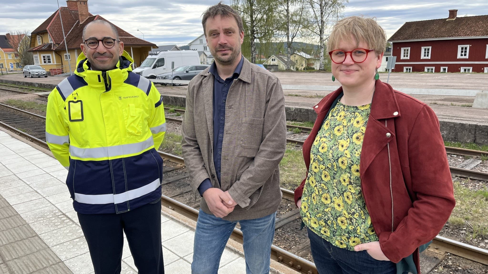 Två män och en kvinna står vid en järnväg på en hållplats