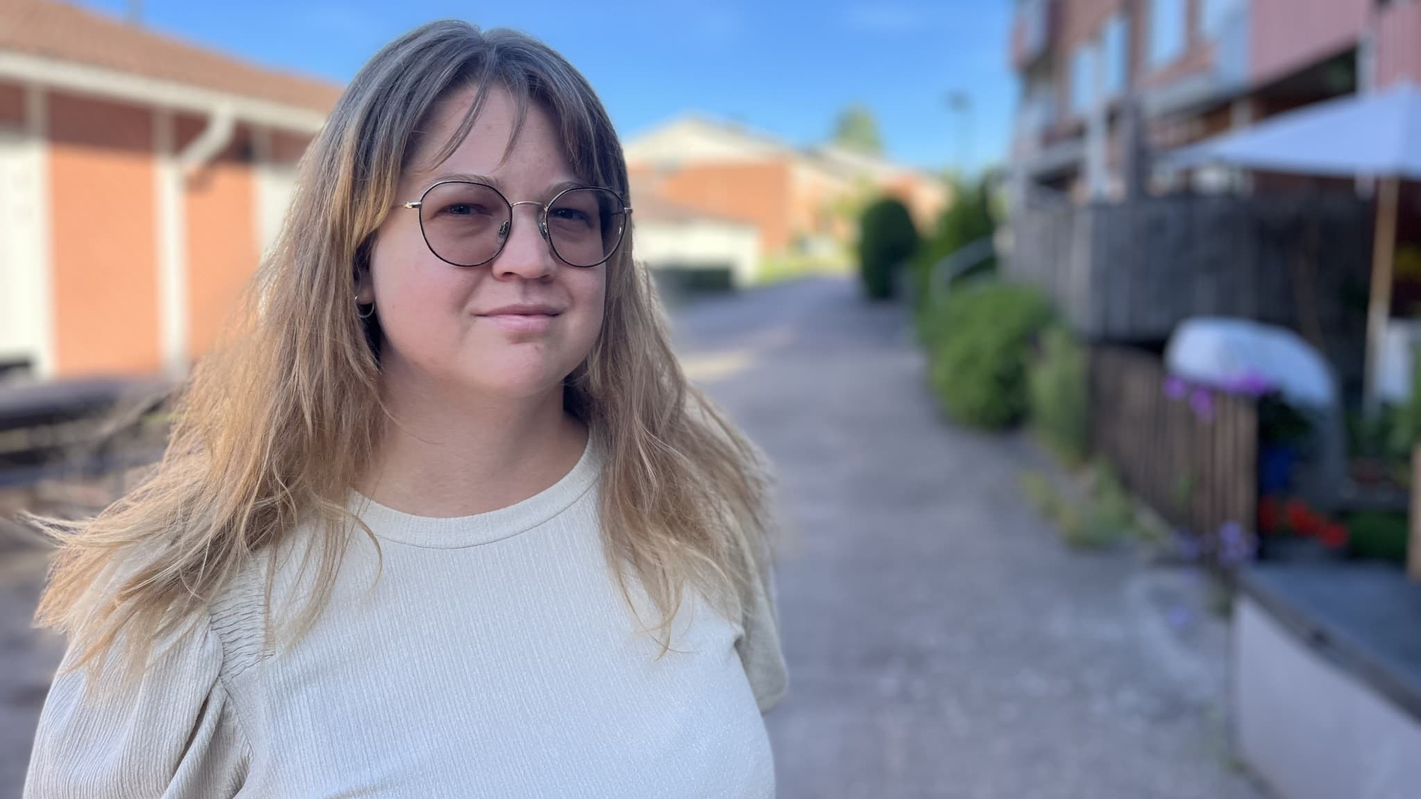 Ida Morales, socialsekreterare, står på en gångväg omgiven av bostadshus i ett bostadsområde. Hon bär en vit tröja och har ljust hår. Soligt väder med klarblå himmel.
