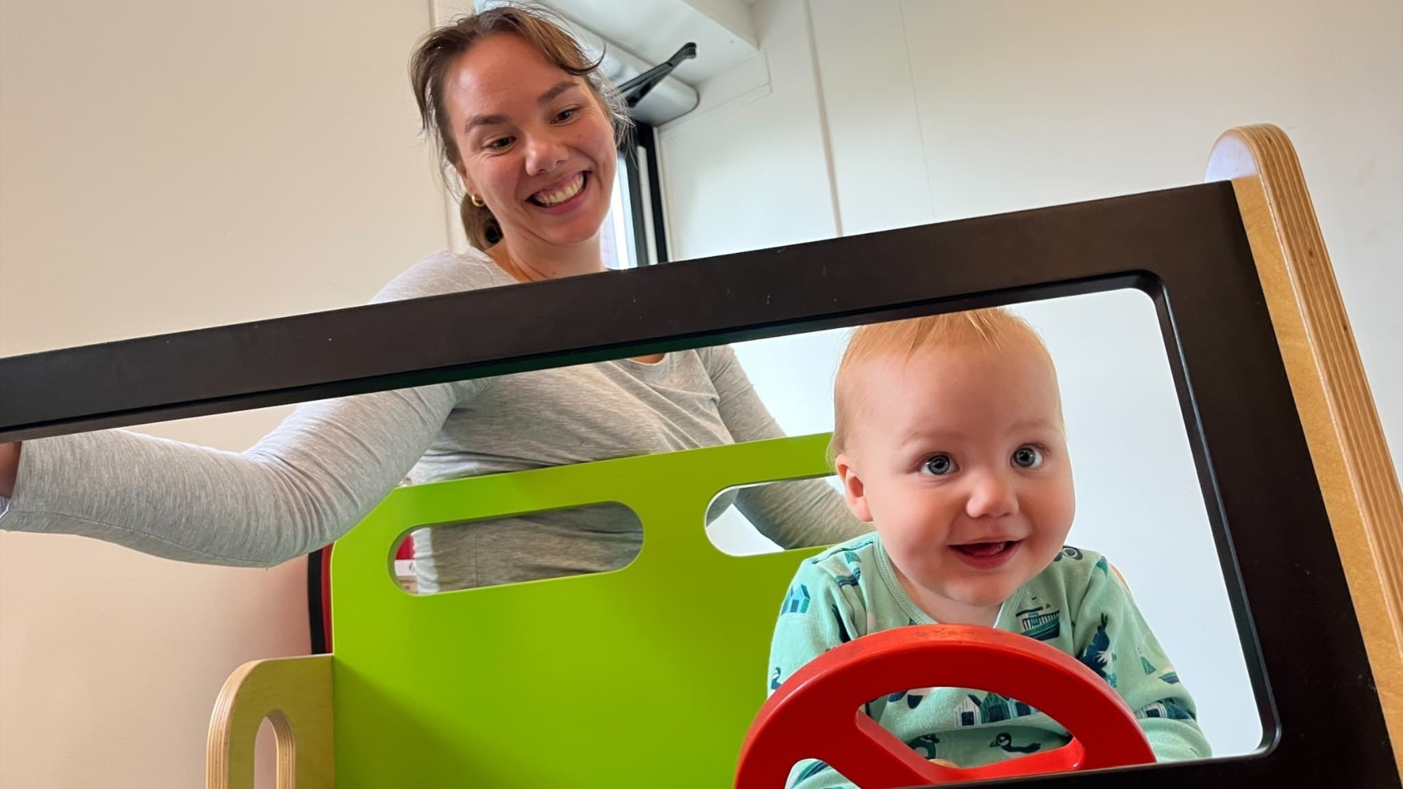 Ylva Engström sitter med sin son Axel i en leksaksbil med grön och röd färg, där Axel håller i ratten. Miljön är inomhus, med ljusa väggar och en modern design.