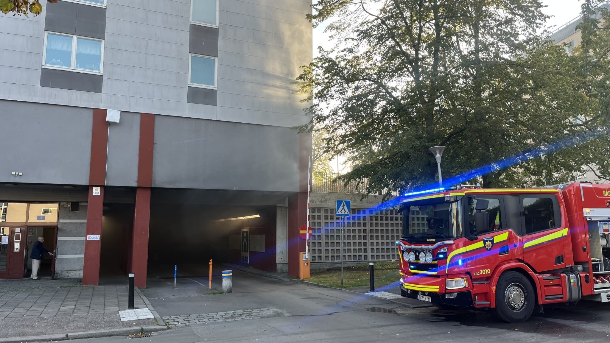 Brand i garage vid flerbostadshus. Rök strömmar ut från byggnaden medan räddningstjänst är på plats med brandbil. Området är avspärrat med gul tejp, och människor står vid sidan.