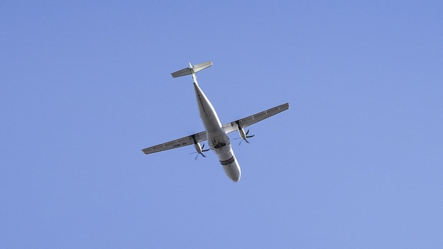 Ett flygplan syns på en klar blå himmel nära Bromma flygplats i Stockholm, med grenar från ett träd och en byggnad i förgrunden. Bilden illustrerar närhet till urban miljö.