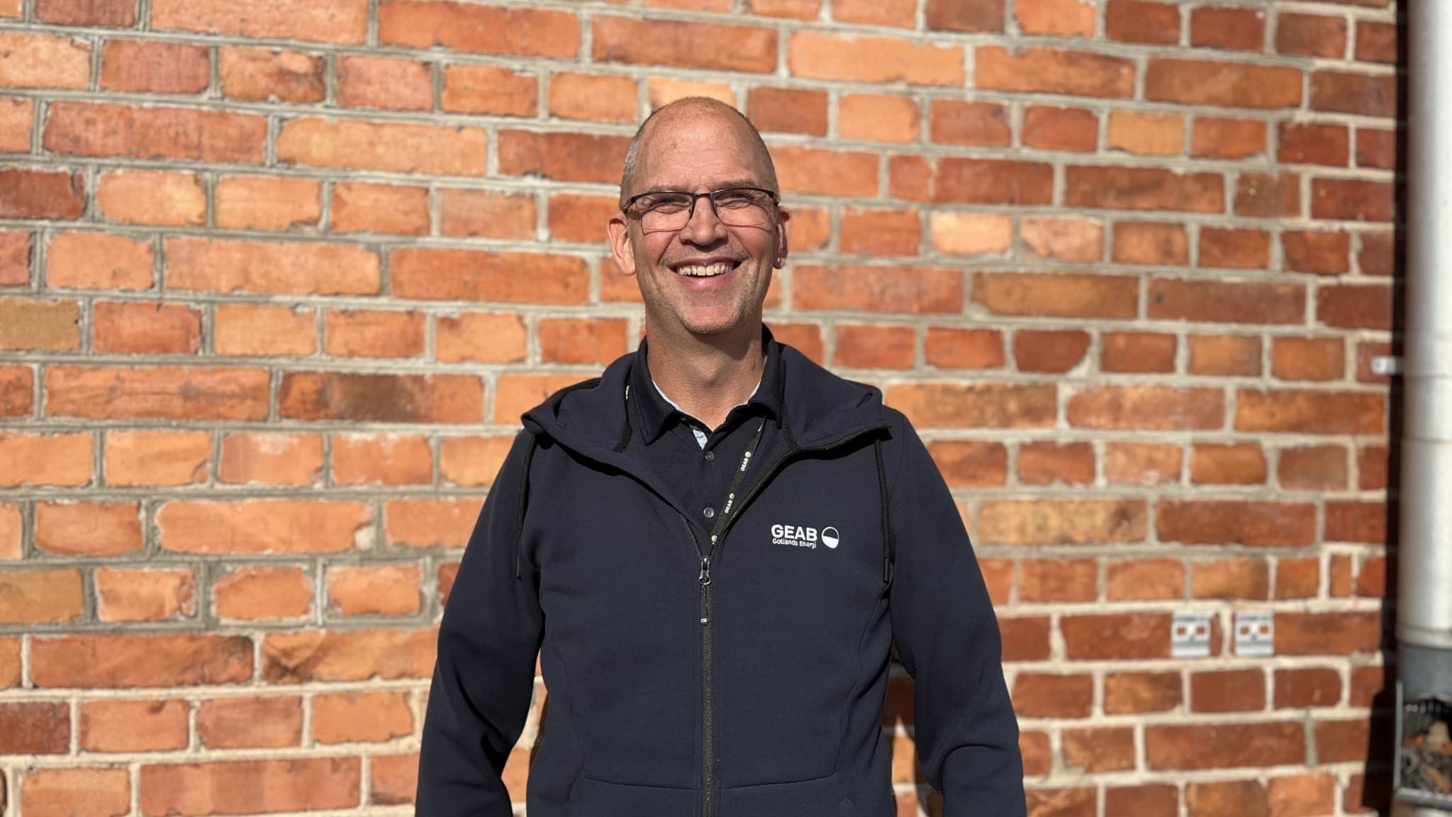 Jonas Hallsten står framför en tegelvägg iklädd en mörkblå arbetsjacka med logotyp. Solen lyser på honom och väggen, vilket skapar en skarp kontrast mellan ljus och skugga.