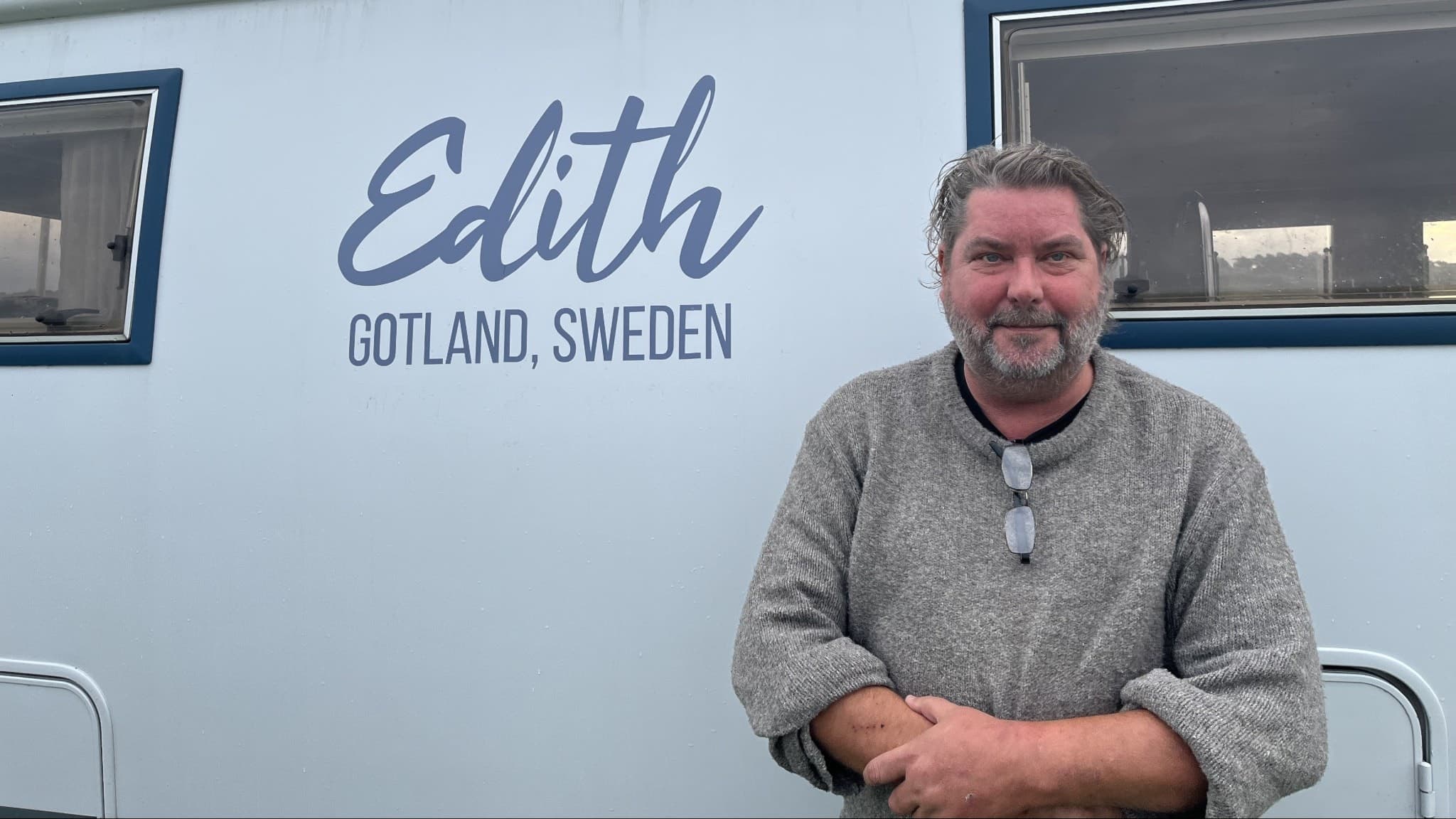 Johan Kurelid står framför en husbil märkt med texten 'Edith Gotland, Sweden'. Han är klädd i en grå tröja och omgivningen visar en modern husbilsdesign med fönster.