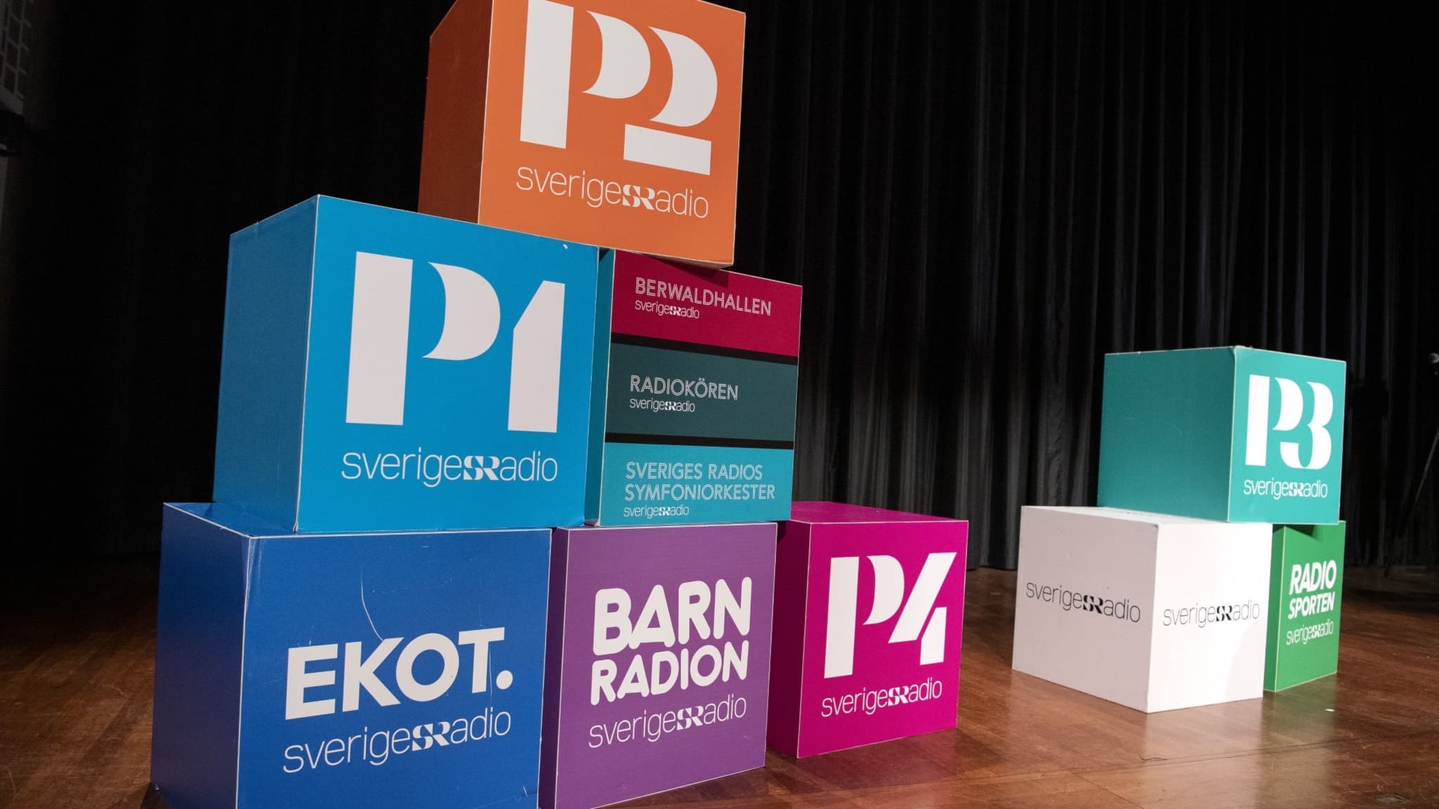 Färgade kuber med logotyper för Sveriges Radios program, inklusive Ekot, Barnradion och P4, står staplade på en scen med mörk bakgrund. En inbjudan att besöka ditt favoritprogram.