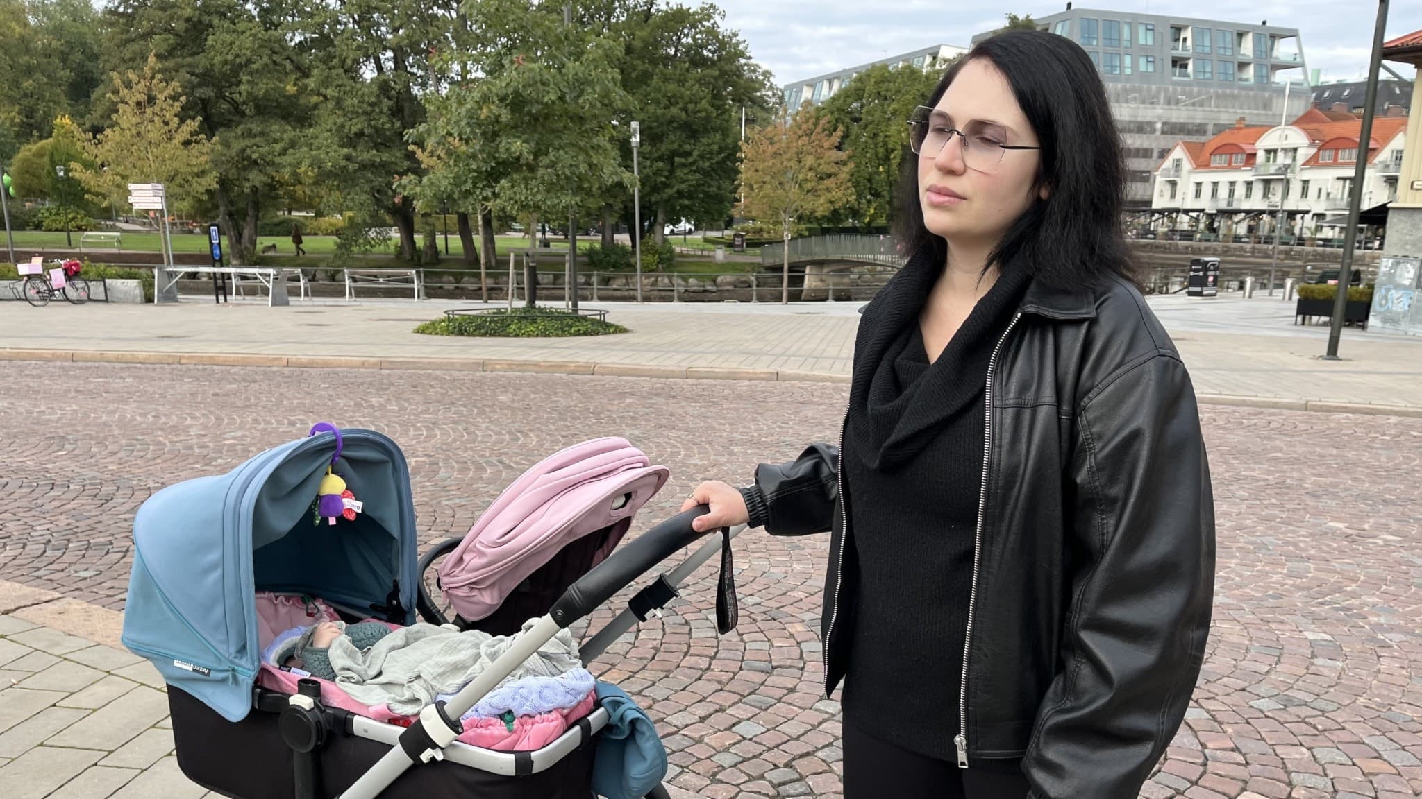 En person i svart jacka står utomhus och håller i en barnvagn med två sittdelar. I bakgrunden syns en park och byggnader i stadsmiljö.