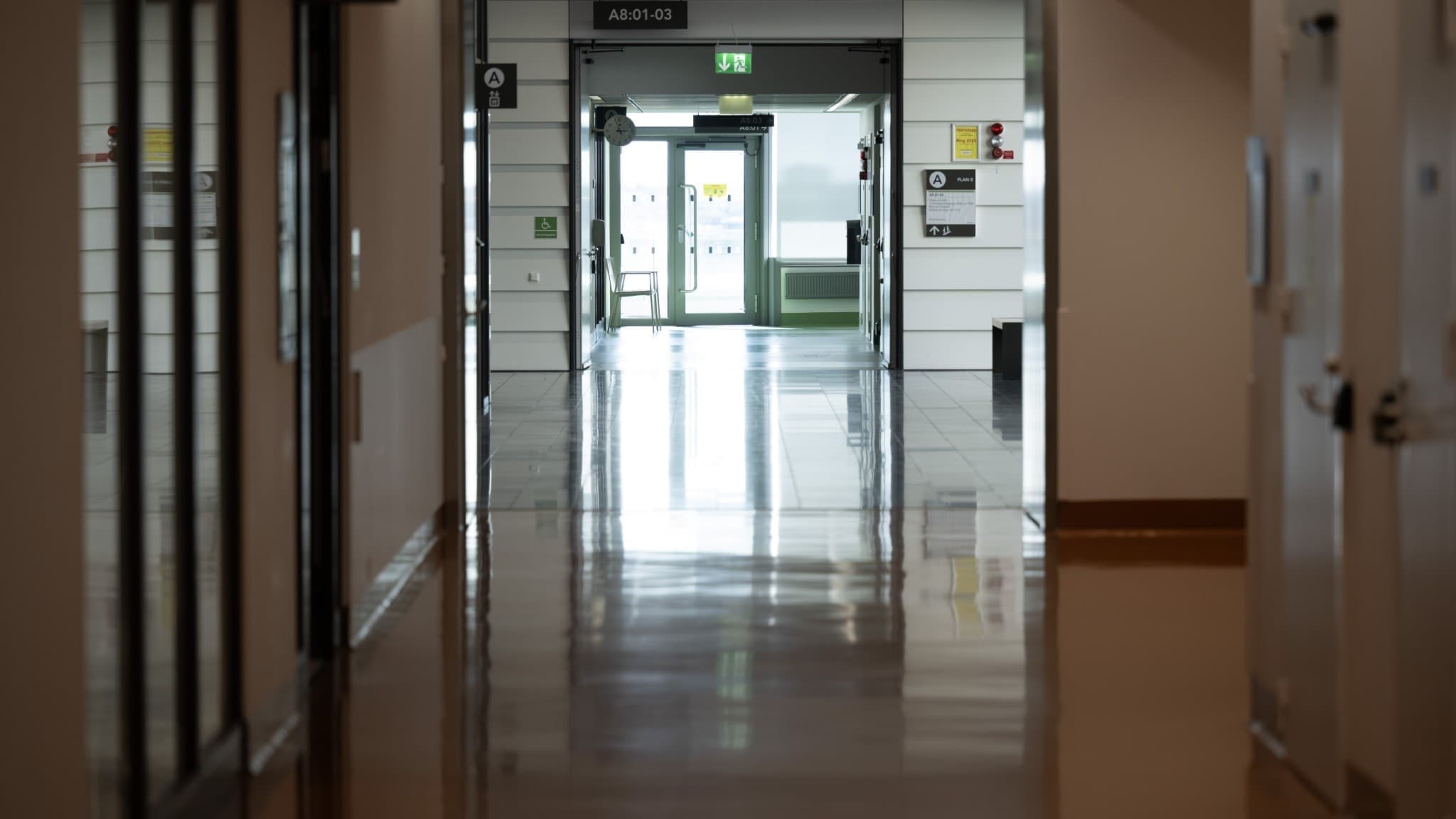 En tom sjukhuskorridor med blankt golv och dörrar på sidorna, fotograferad på Karolinska universitetssjukhuset i Solna. Nödutgångsskylt syns i bakgrunden.