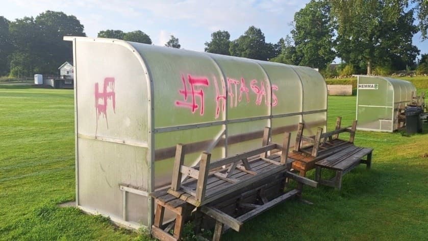 Avbytarbås tillhörande Horreds IF på en fotbollsplan, nedklottrat med graffiti. Gräsplan och träd syns i bakgrunden under en solig dag.