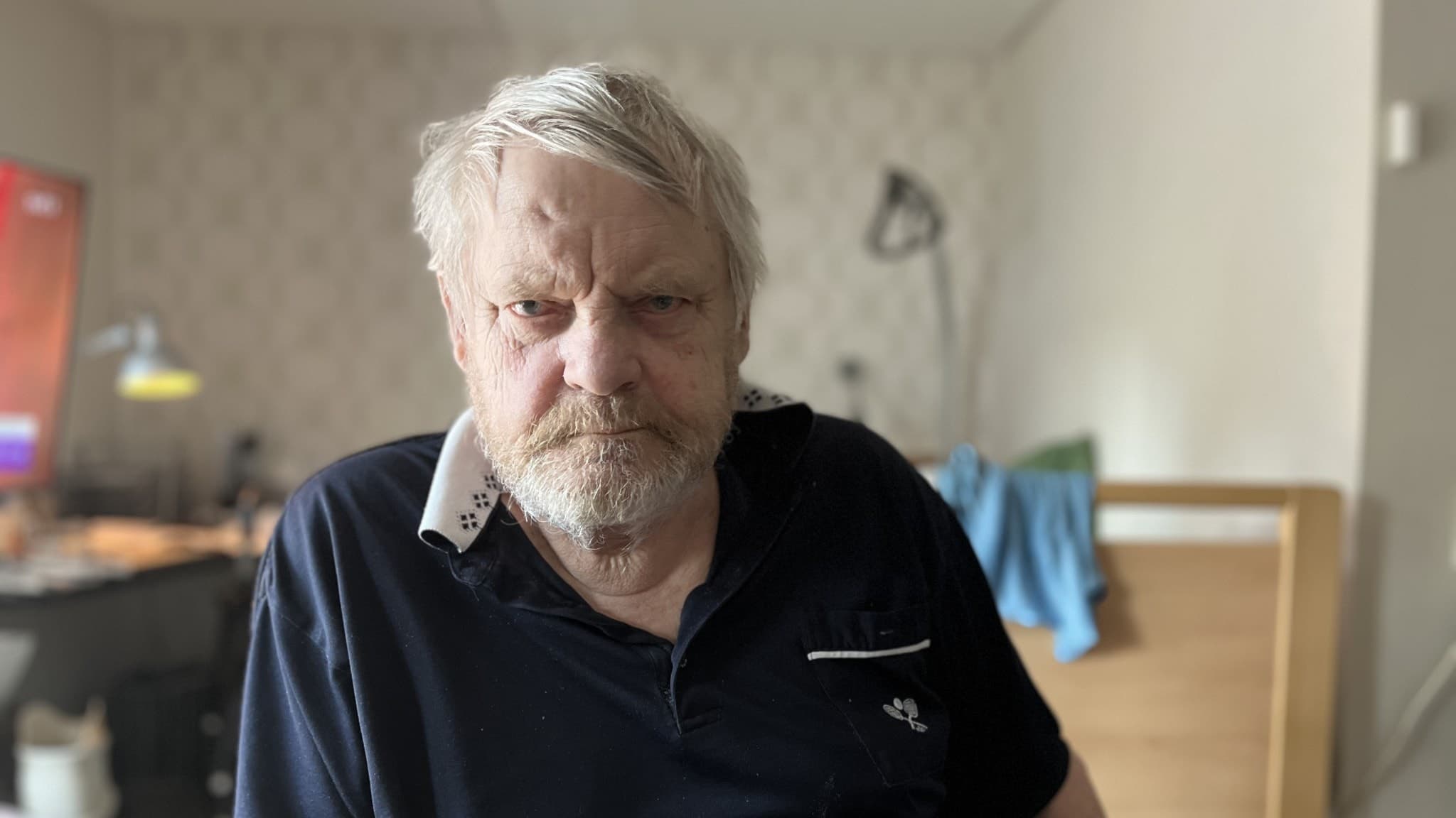 En äldre man, Kjell Åke Bergkvist, sitter i ett rum med säng och skrivbordslampa i bakgrunden. Han har grått hår och är klädd i en mörkblå tröja. Miljön ser ut som ett vårdboende.