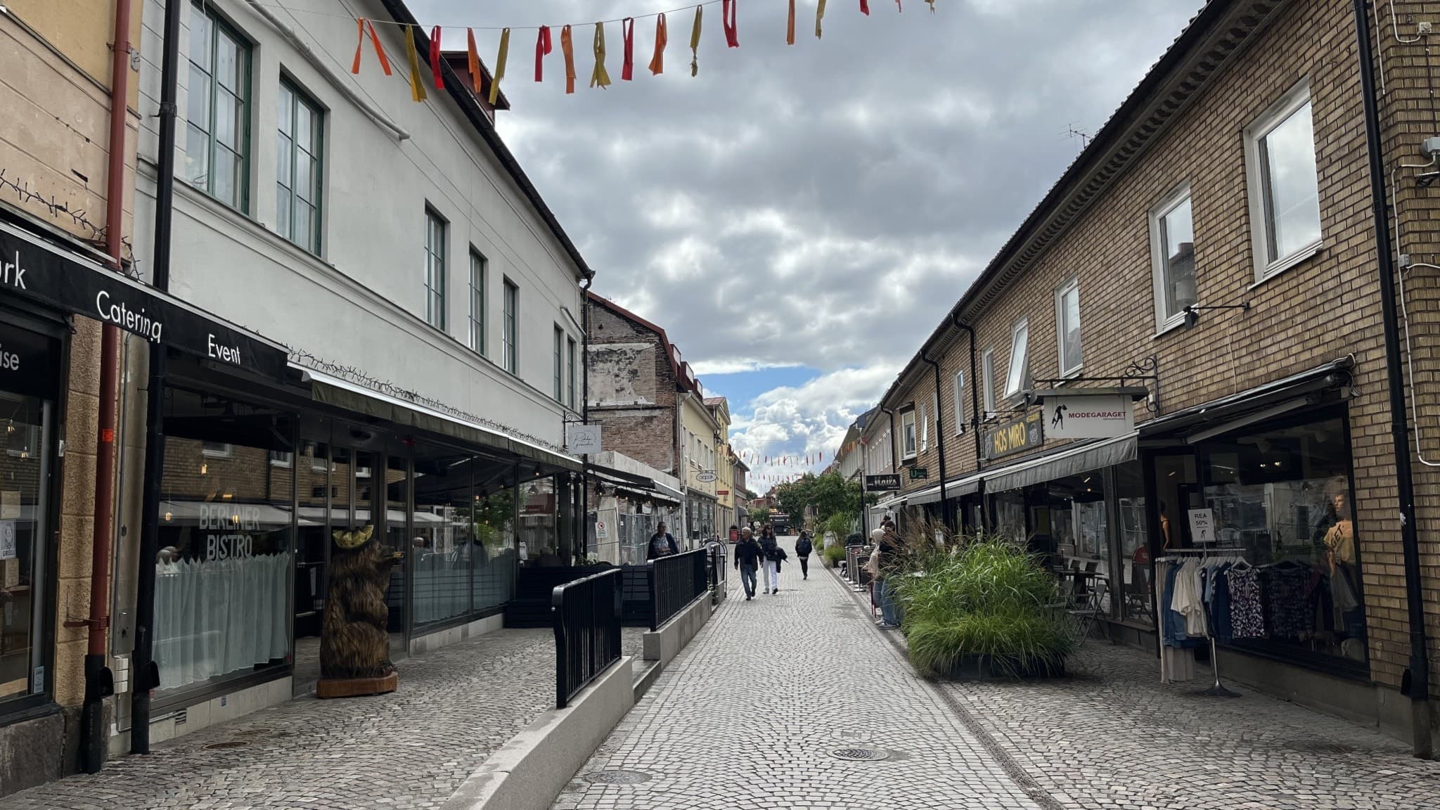 Storgatan i Ulricehamn med människor som promenerar längs en stenlagd gata omgiven av butiker och caféer. Färgglada vimplar hänger över gatan under en molnig himmel.