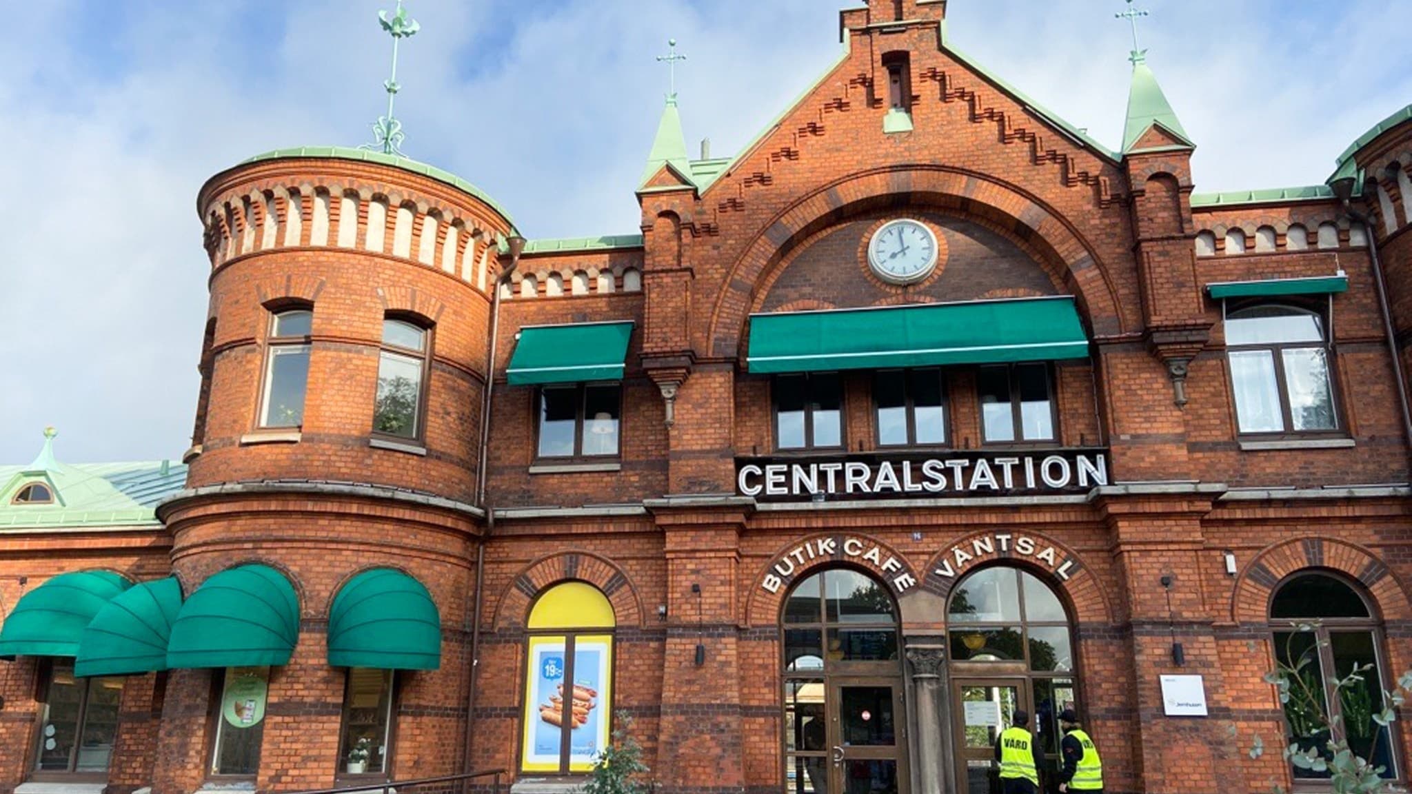 Borås centralstation i röd tegelbyggnad med gröna markiser och texten 'Centralstation' synlig på fasaden. Två personer står utanför ingången. Tågtrafikens höga kostnader diskuteras.