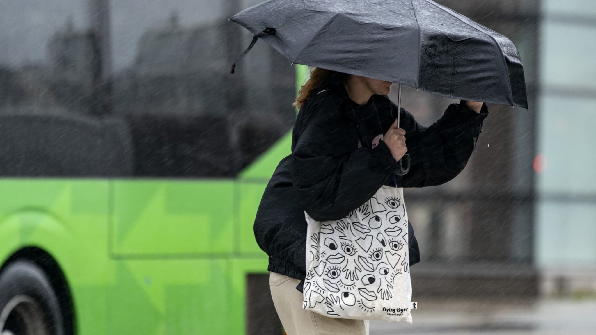 En kvinna med svart paraply kämpar mot kraftiga vindar vid Malmö C, medan en grön buss passerar i bakgrunden. Sydvästra Sverige drabbas av regn och stormvindar enligt SMHI:s varning.