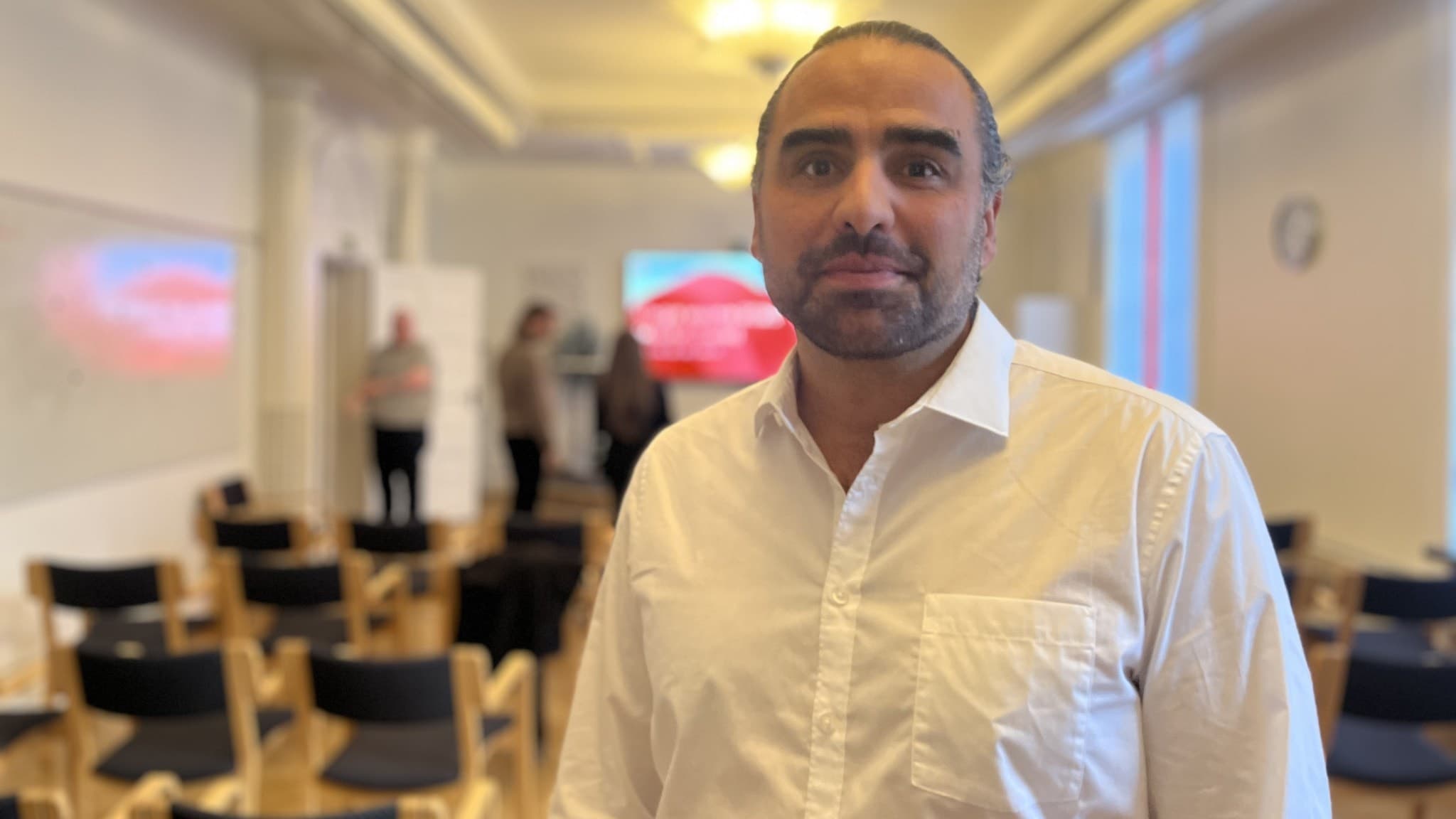 Bostadsutredare Ragnar Bengtsson står i ett konferensrum med stolar och en stor skärm i bakgrunden. Flera personer samtalar längre bak i rummet.