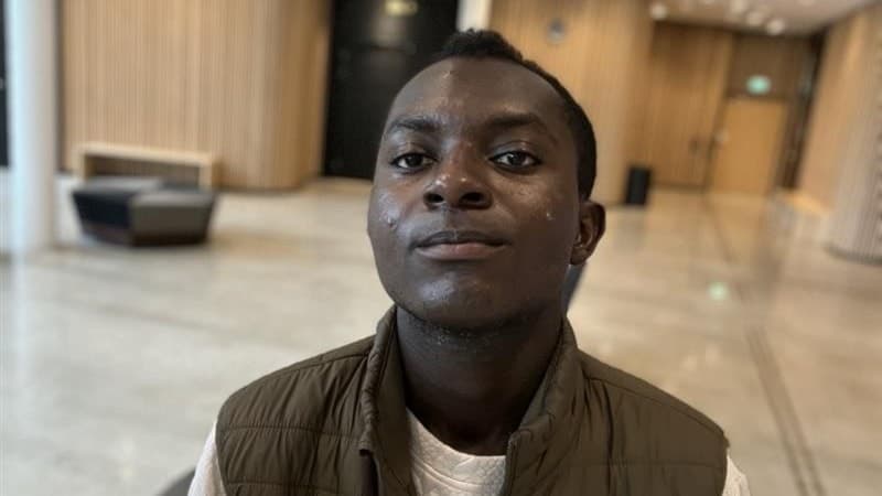 En man med mörk hudton klädd i vit tröja och grön väst står i ett ljust rum med träpaneler och möbler. Han har blivit hotad med kniv av sin granne efter att ha klagat på störningar.