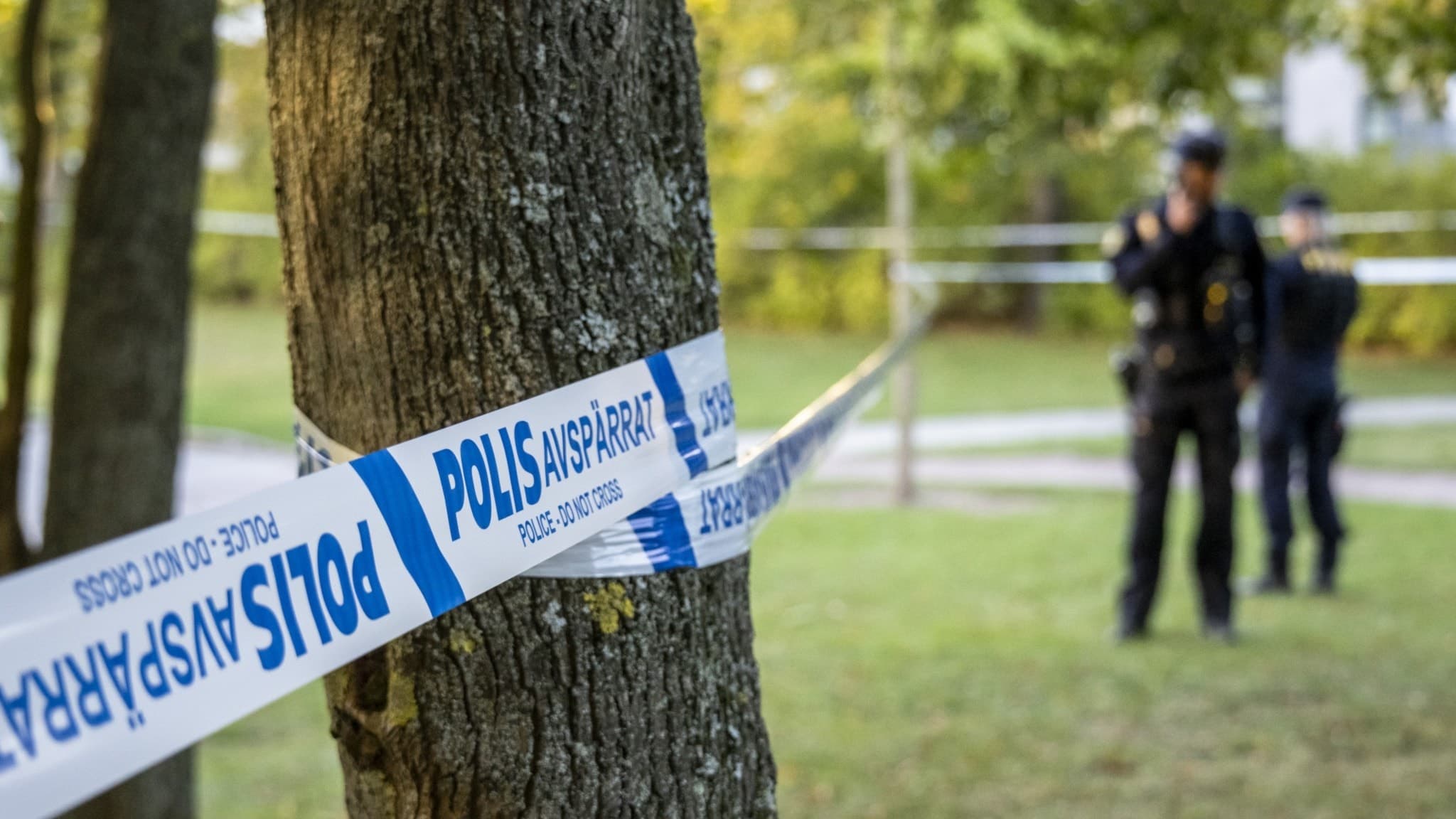 Polisavspärrning kring ett grönområde i Malmö efter ett misstänkt mordförsök. En polis syns i bakgrunden nära ett träd. Händelsen utreds och en skadad person har förts till sjukhus.