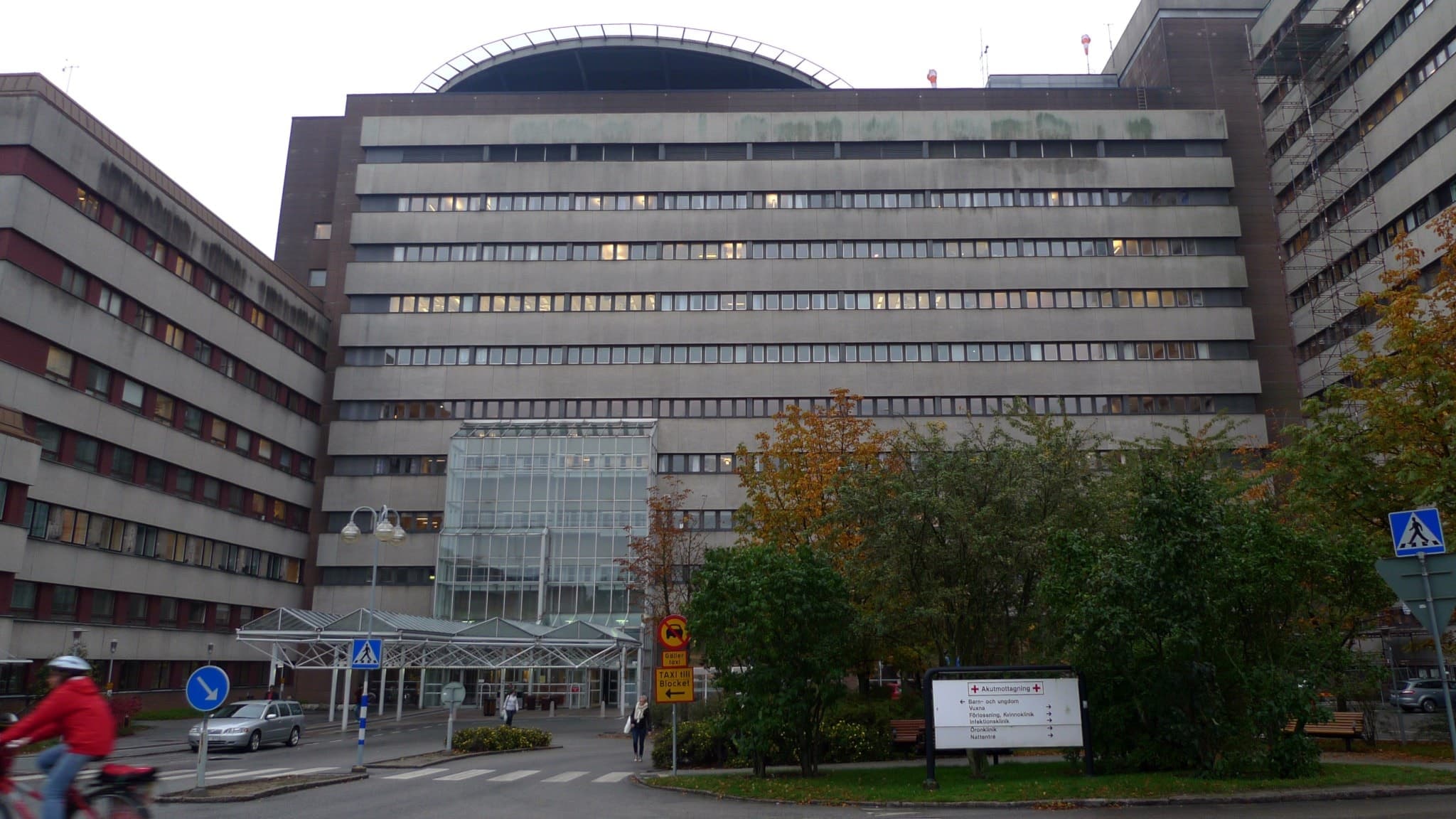 Skånes Universitetssjukhus i Lund, med huvudblocket och helikopterplatta. Byggnaden omges av träd och en väg där cyklister och skyltar syns i förgrunden.