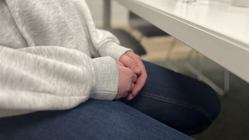 En person i grå hoodie och jeans sitter vid ett vitt bord med händerna knäppta. På bordet står ett glas vatten och en vit mugg. Bakgrunden består av ett ljusgrått rum med enkla möbler.