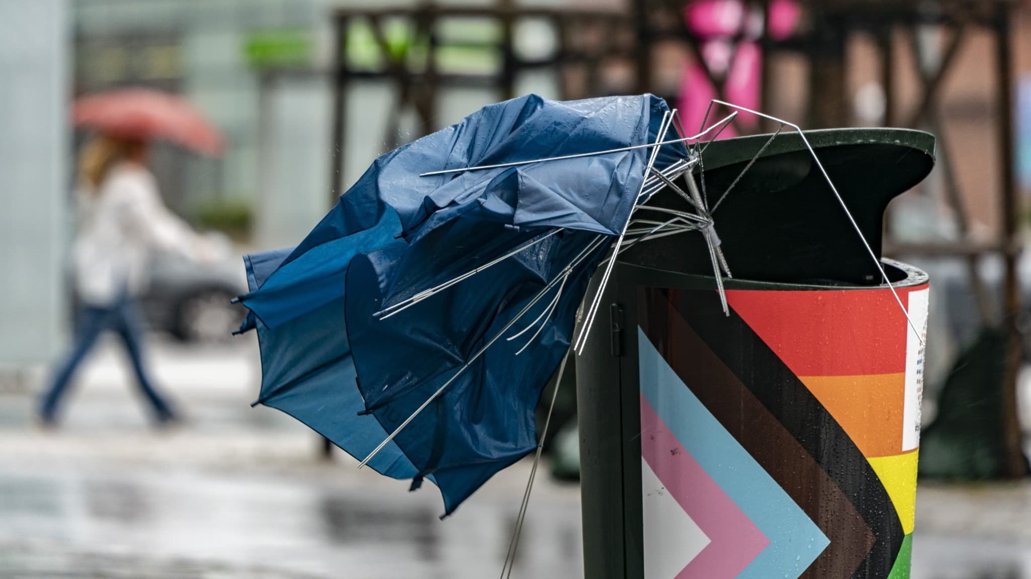 Ett trasigt blått paraply står fastklämt i en papperskorg på en regnig gata i Malmö. I bakgrunden syns en person med paraply och suddiga byggnader. Stormbyar och busväder präglar södra Sverige.